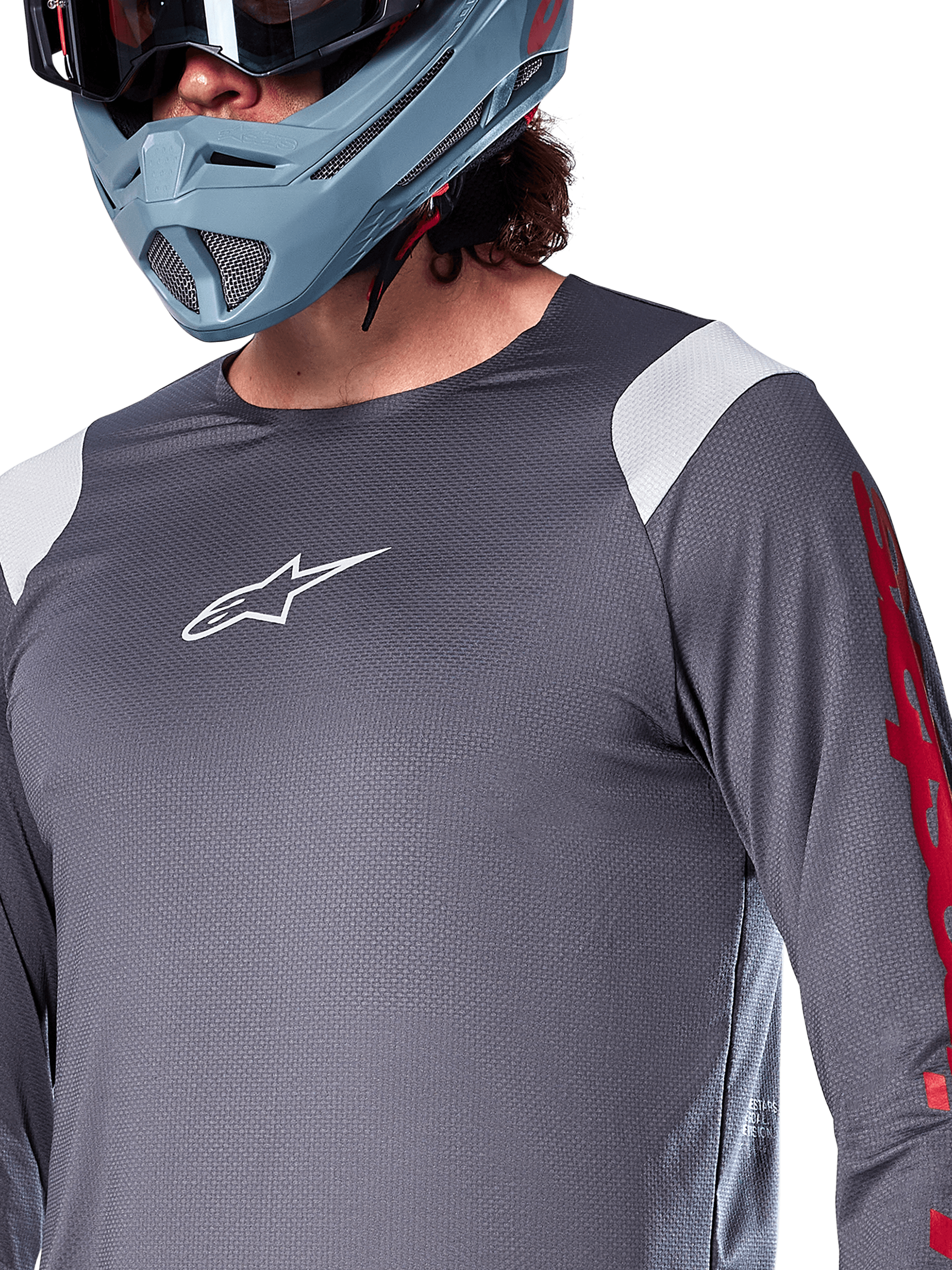 Alpinestars A-Supra Astar Jersey met lange mouwen, donkergrijs met witte schouderpanelen en rode graphics op de mouwen, gedragen met een licht blauwgrijze full-face helm, performance mountainbikekleding