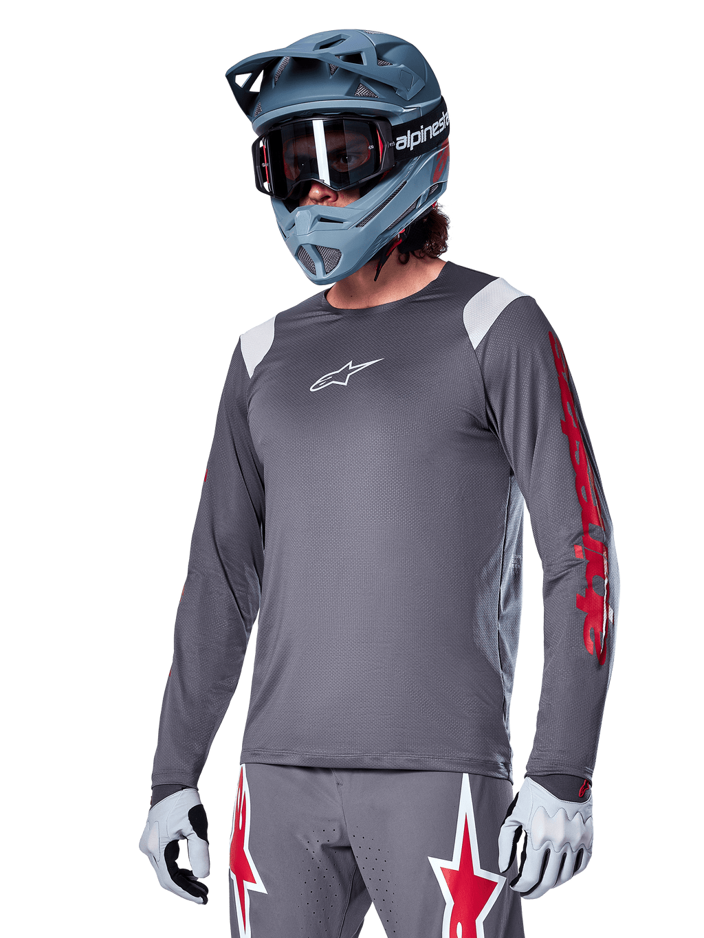 Alpinestars A-Supra Astar jersey met lange mouwen, MTB mountainbike jersey, donkergrijs met rode en witte accenten, sportieve pasvorm, gedragen met bijpassende grijze broek met rode en witte sterren-graphics, complete off-road fietsoutfit met full-face helm en handschoenen