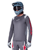 Alpinestars A-Supra Astar jersey met lange mouwen, MTB mountainbike jersey, donkergrijs met rode en witte accenten, sportieve pasvorm, gedragen met bijpassende grijze broek met rode en witte sterren-graphics, complete off-road fietsoutfit met full-face helm en handschoenen