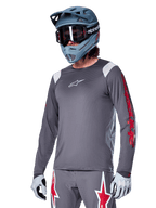 Alpinestars A-Supra Astar jersey met lange mouwen, MTB mountainbike jersey, donkergrijs met rode en witte accenten, sportieve pasvorm, gedragen met bijpassende grijze broek met rode en witte sterren-graphics, complete off-road fietsoutfit met full-face helm en handschoenen