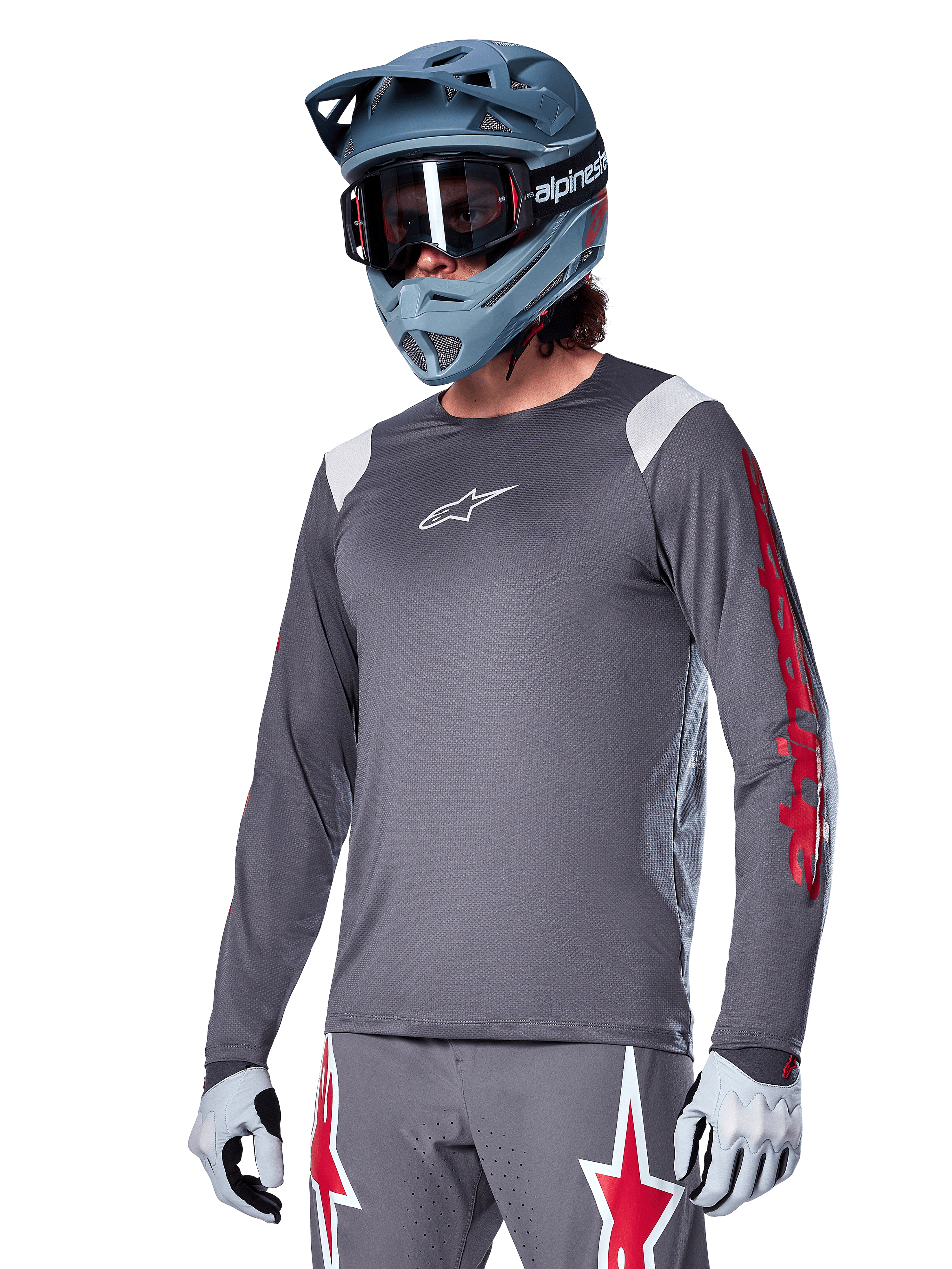 Alpinestars A-Supra Astar jersey met lange mouwen, MTB mountainbike jersey, donkergrijs met rode en witte accenten, sportieve pasvorm, gedragen met bijpassende grijze broek met rode en witte sterren-graphics, complete off-road fietsoutfit met full-face helm en handschoenen