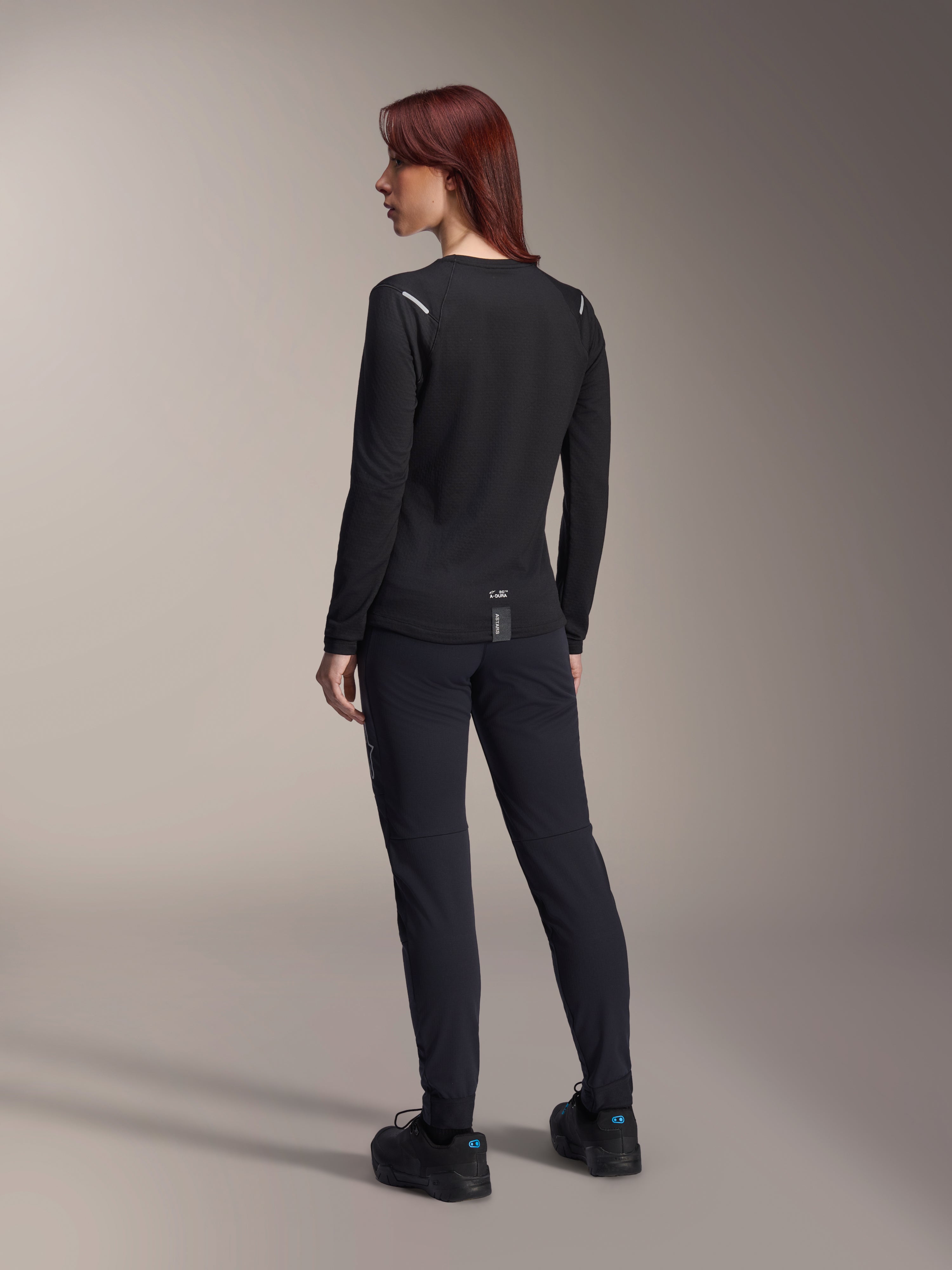 Alpinestars Stella A-Dura Thermal Jersey voor dames, MTB-shirt met lange mouwen in het zwart, achteraanzicht met reflecterende schouderaccenten en verlengde drop tail, gedragen met zwarte broek en schoenen, performance kleding voor mountainbiken