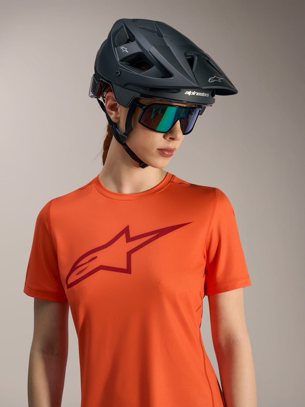 Maillot Alpinestars Stella A-Dura Ride orange avec logo rouge, casque VTT noir et lunettes irisées.