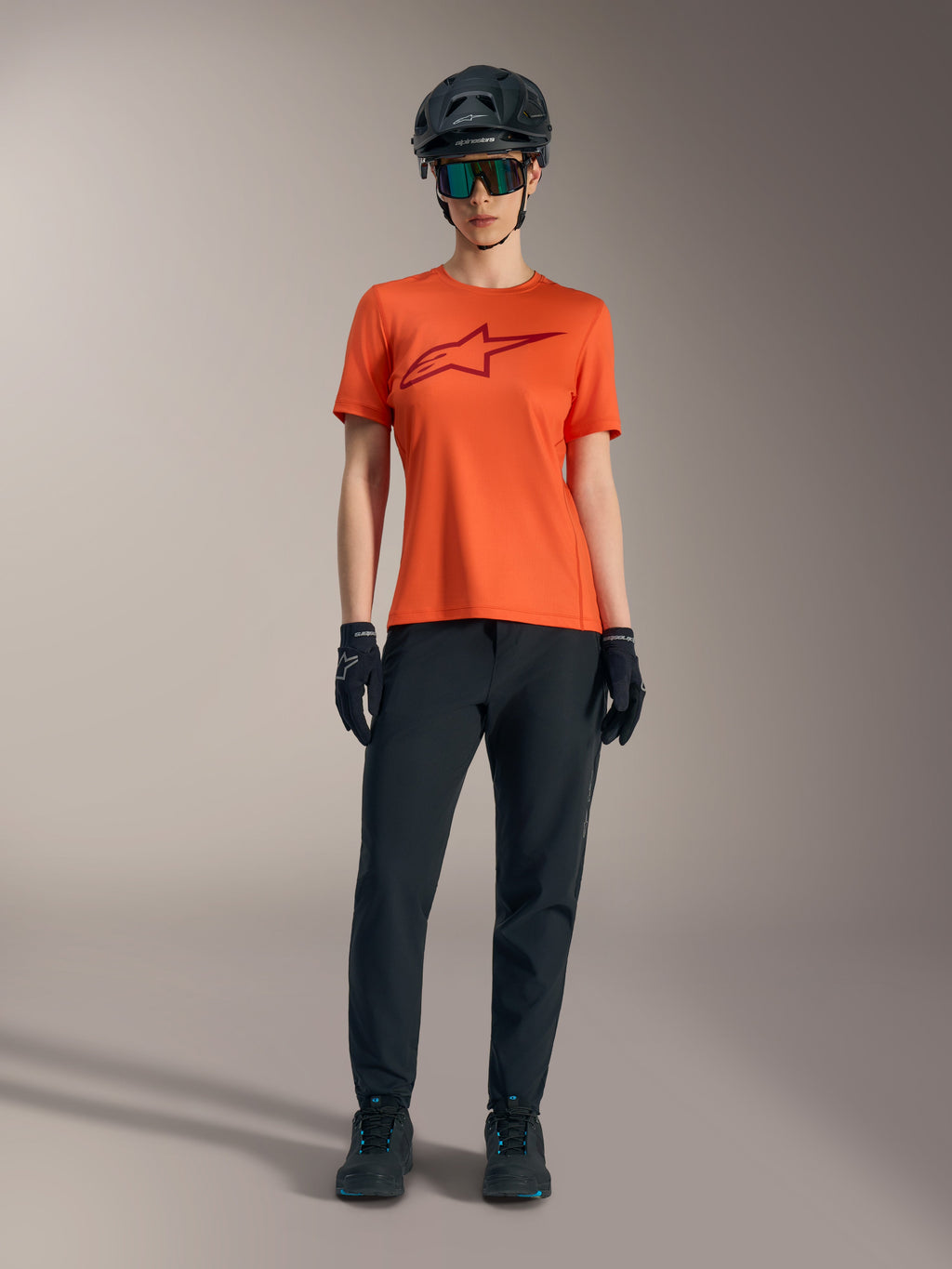 Maillot femme Stella A-Dura Ride orange à manches courtes, logo bordeaux, avec équipement VTT, casque et gants noirs.