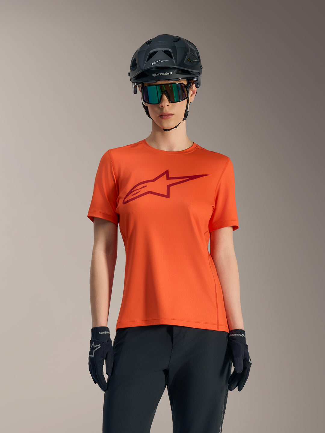 Maillot orange Stella A-Dura Ride femme à manches courtes, logo rouge, avec casque de VTT et lunettes noirs.