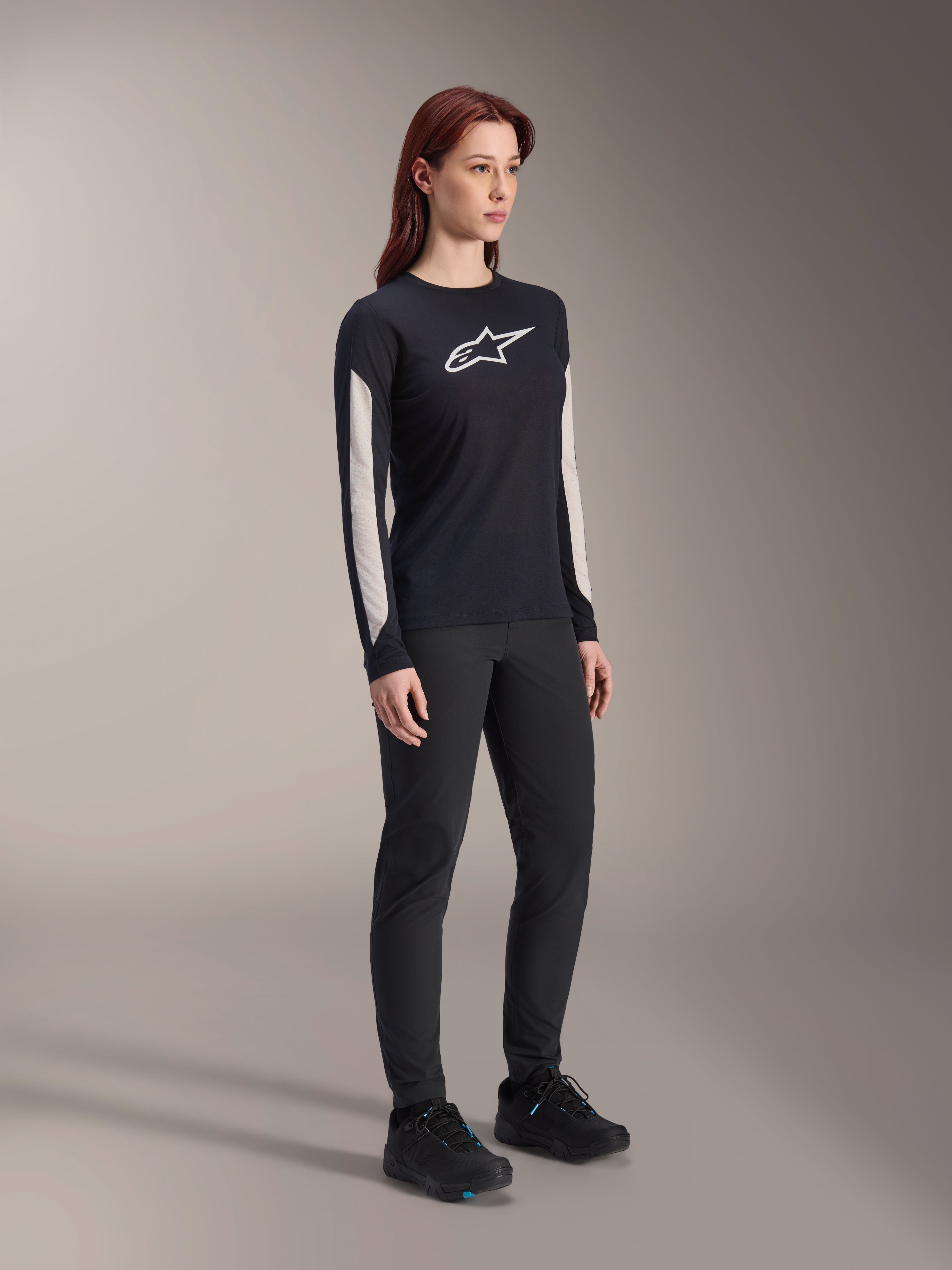 Femme portant une tenue de VTT Alpinestars, maillot Stella A-Dura Elite noir avec logo blanc sur la poitrine et empiècements aux manches, pantalon Stella A-Dura noir, chaussures de cyclisme noires avec accents bleus, tenue de performance complète pour le VTT