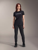 Modèle portant le maillot à manches courtes Alpinestars Stella A-Dura Dri Switch pour femme en noir, avec logo blanc sur la poitrine, associé à un pantalon technique noir et des chaussures de sport noires, photo de studio en pied