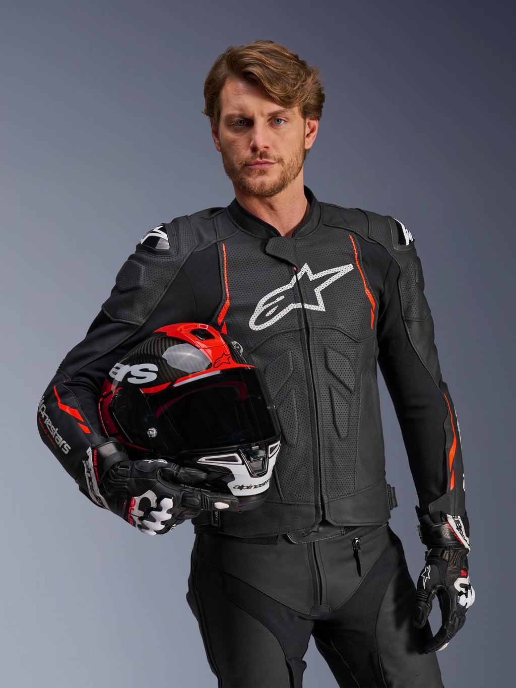 Veste en cuir Alpinestars Dusk Airflow, noire avec accents rouge fluo, portée par un mannequin homme tenant un casque de moto rouge et noir, dotée de cuir perforé pour une ventilation maximale et de logos Alpinestars blancs, équipement de moto sport