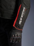 Alpinestars T-Sps Air V2 Jacke, Nahaufnahme des Ärmels in Schwarz-Rot-Fluo, mit weißen Alpinestars und T-SPS AIR Logos, leuchtend rotem fluoreszierendem Streifen, getragen mit einem schwarzen Motorrad-Schutzhandschuh mit Carbon-Knöchelschutz