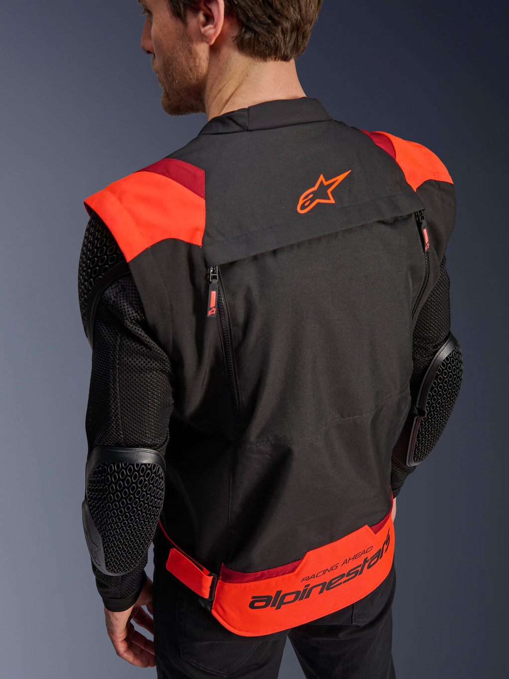 Alpinestars T-Stunt Air jas, zwart, fluorood en donkerrood, achteraanzicht op een model, toont mesh mouwen met elleboogbeschermers met honingraatstructuur, Alpinestars-sterlogo op de bovenrug en opvallende branding op de onderrug met 'RACING AHEAD' tekst, modulair sportief ontwerp voor motorrijders.