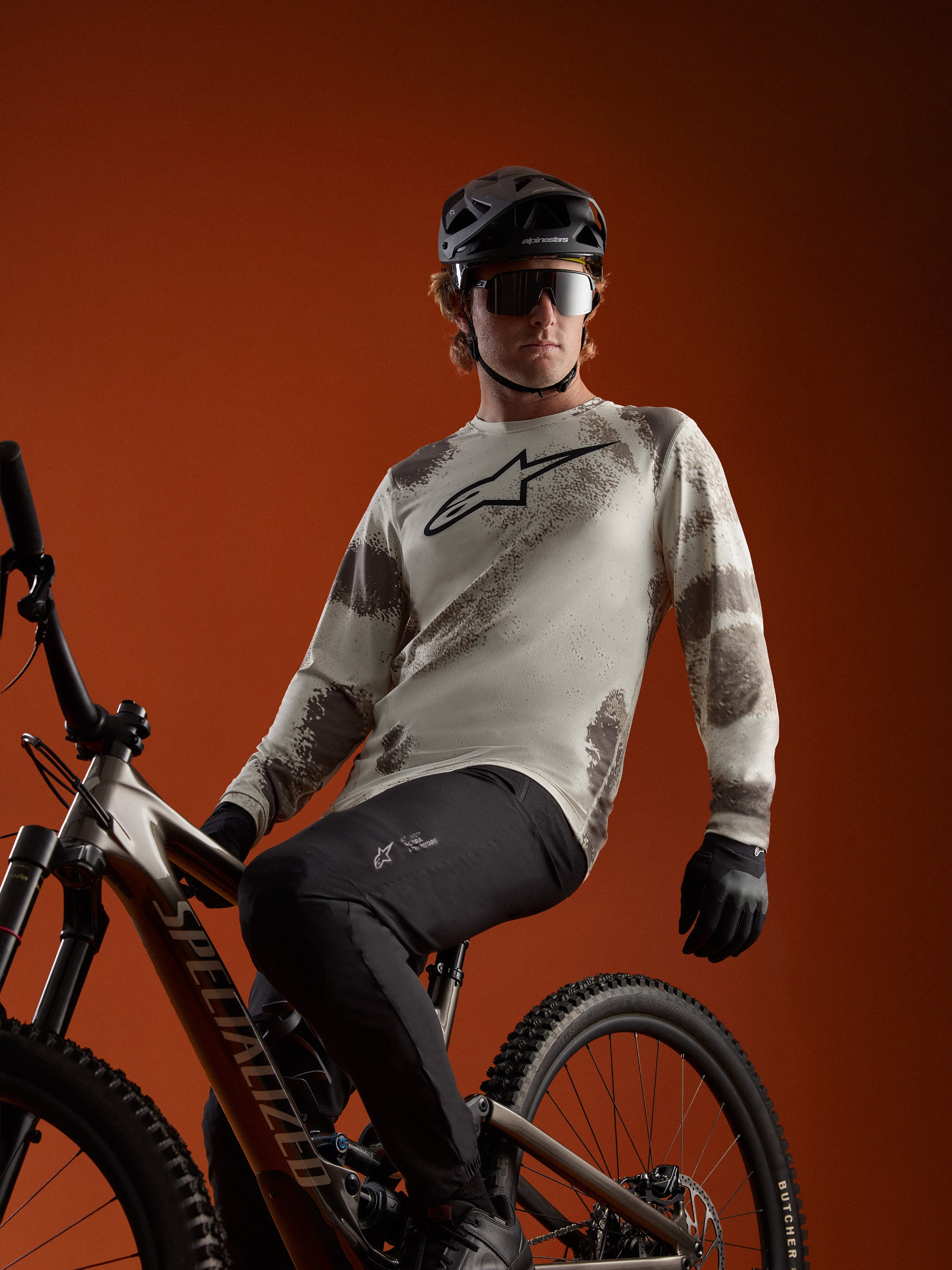 Maillot A-Dura Ride à manches longues Sand Camo avec logo Alpinestars, porté par un cycliste sur un VTT.
