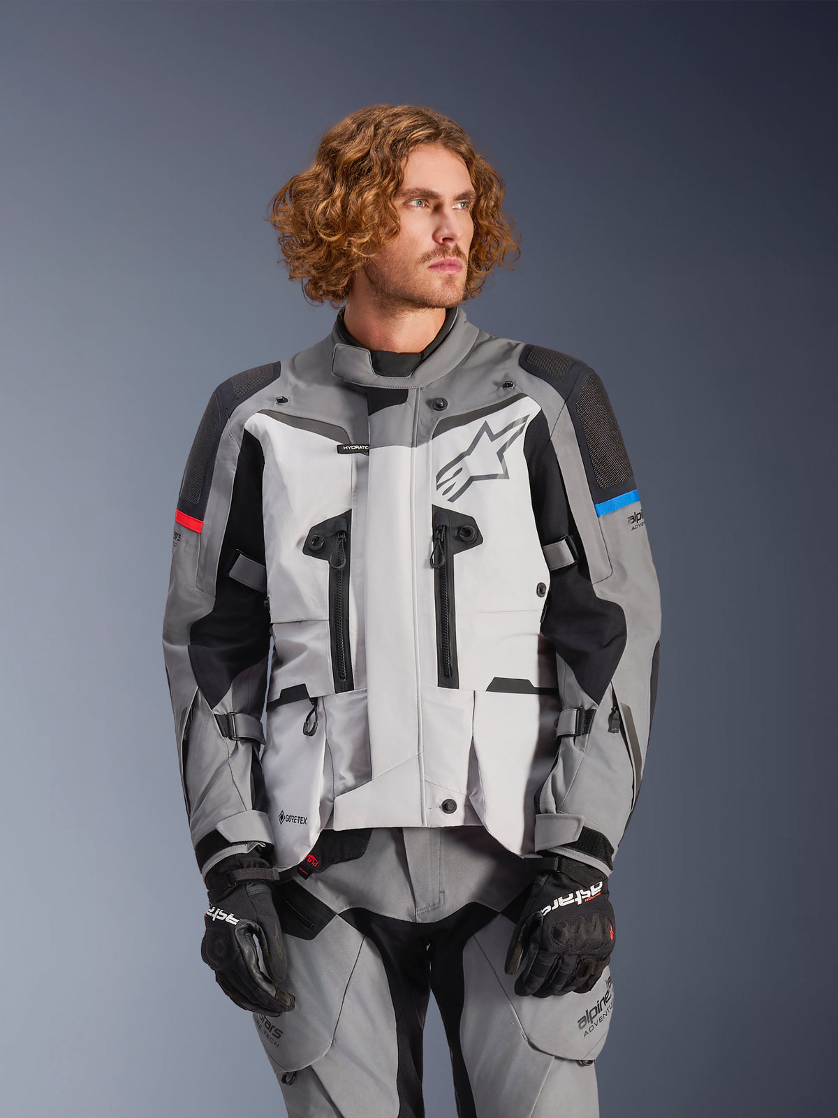 Alpinestars Boulder Gore-Tex, veste de moto touring mixte, grise et noire avec accents rouges et bleus, imperméable et respirante, portée avec pantalon et gants assortis