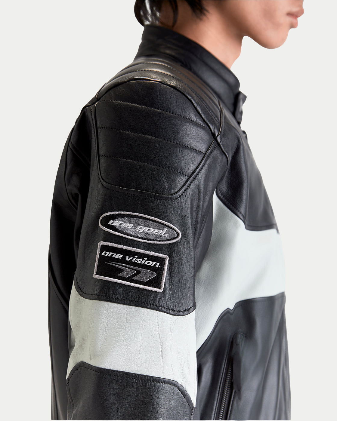 Alpinestars Revern Leather Jacket, black and off-white motorcycle leather jacket, close-up view of the sleeve showing quilted shoulder panels and embroidered 'one goal, one vision' patches worn by a model