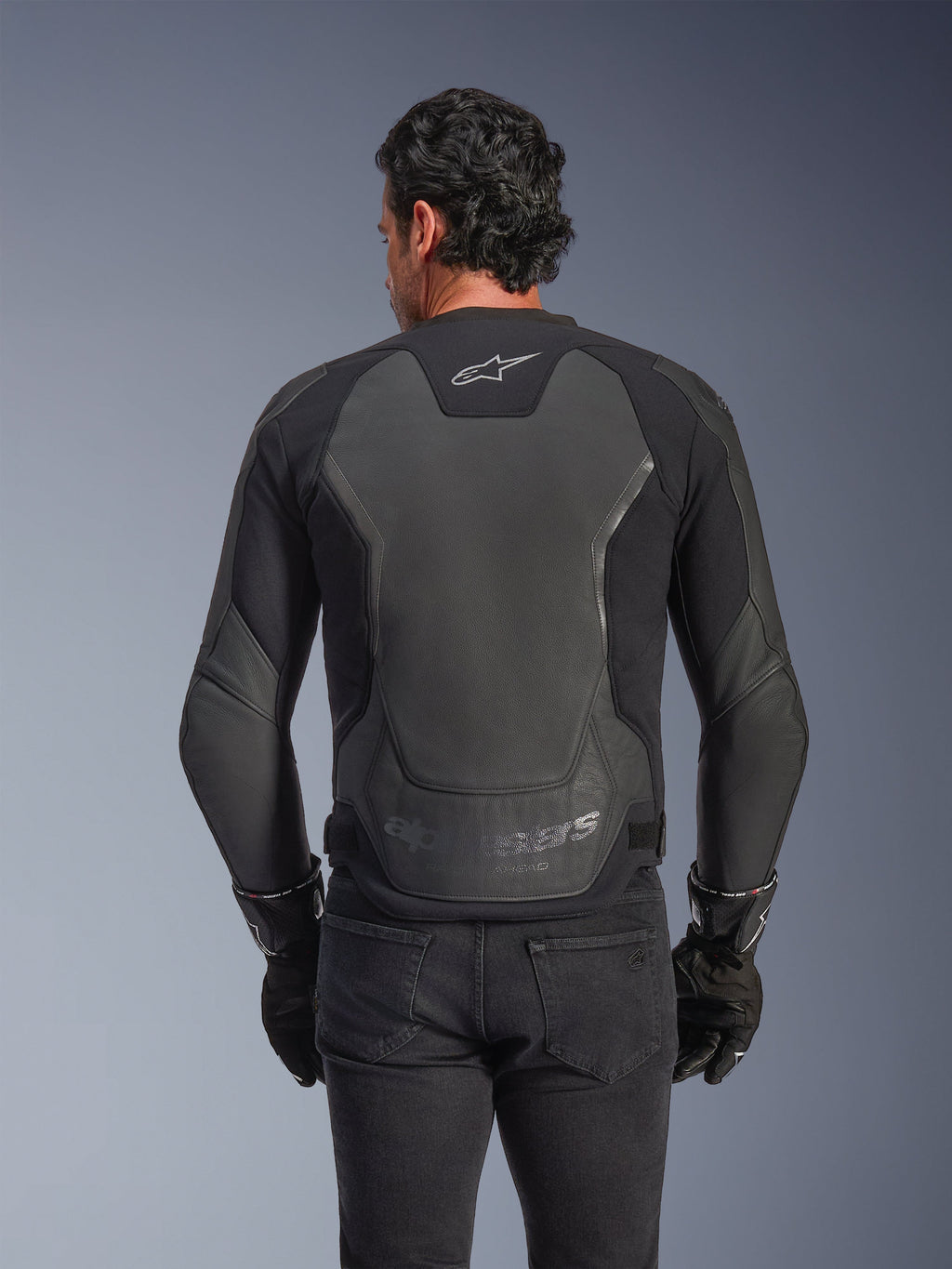 Veste en cuir Alpinestars GP Force V2, noire, vue arrière d'un homme portant la veste avec des gants noirs et un jean, dotée de larges panneaux de protection en cuir dans le dos, le logo Alpinestars sur le haut du dos et un marquage embossé au bas de la taille, équipement de moto d'inspiration sportive