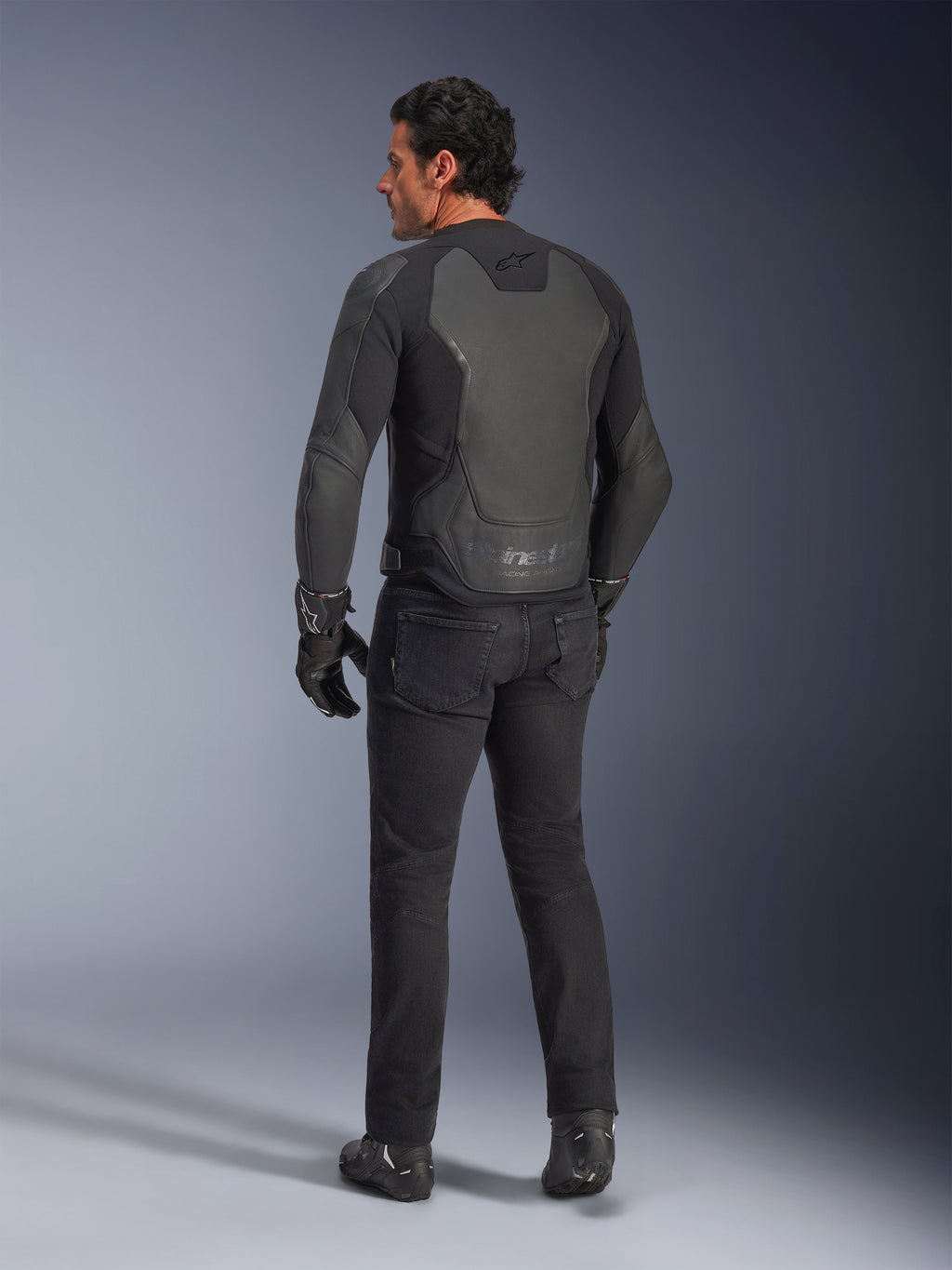 Vue arrière d'un mannequin portant la veste en cuir Alpinestars GP Force V2 en coloris Black Black, dotée d'une construction multi-panneaux et d'une coupe sport, associée à un pantalon, des gants et des bottes de moto noirs pour un look complet inspiré de la course moto.