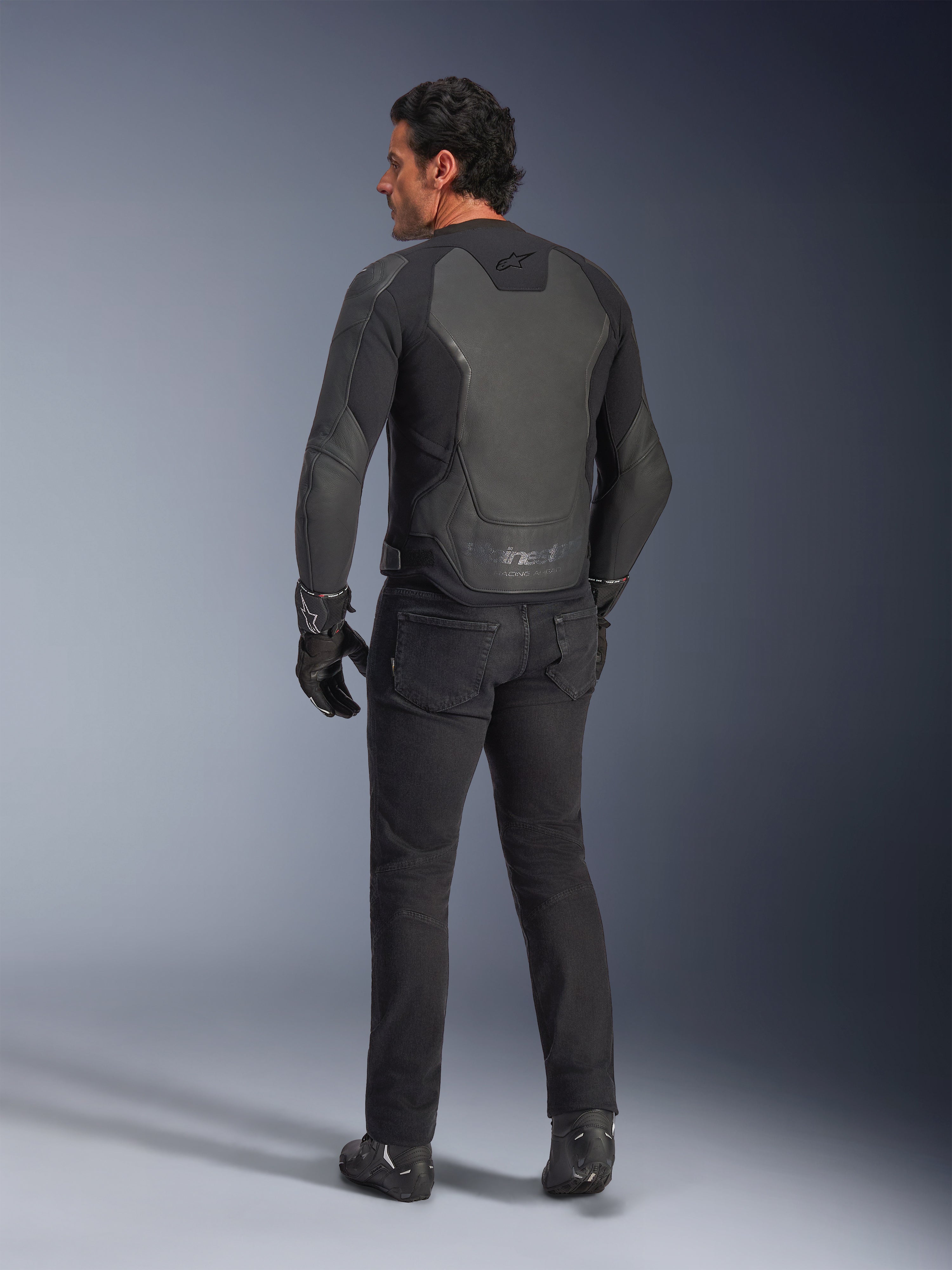 Vue arrière d'un mannequin portant la veste en cuir Alpinestars GP Force V2 en coloris Black Black, dotée d'une construction multi-panneaux et d'une coupe sport, associée à un pantalon, des gants et des bottes de moto noirs pour un look complet inspiré de la course moto.