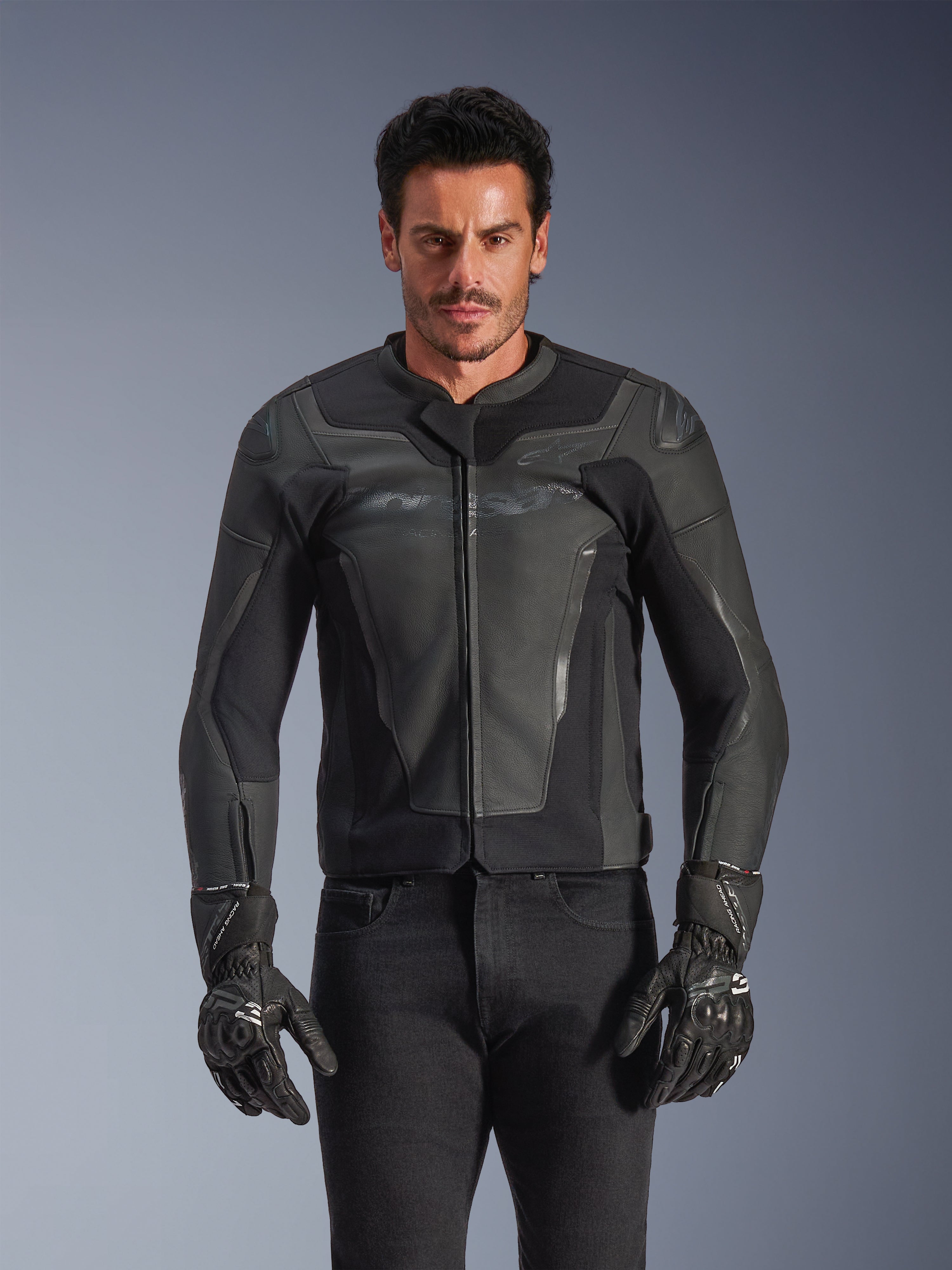 Alpinestars GP Force V2 Leather Jacket, noire et gris foncé, veste de course de moto, homme portant un design ajusté en cuir avec fermeture éclair frontale, associé à des gants texturés noirs, équipement de protection moderne, construction en cuir haut de gamme