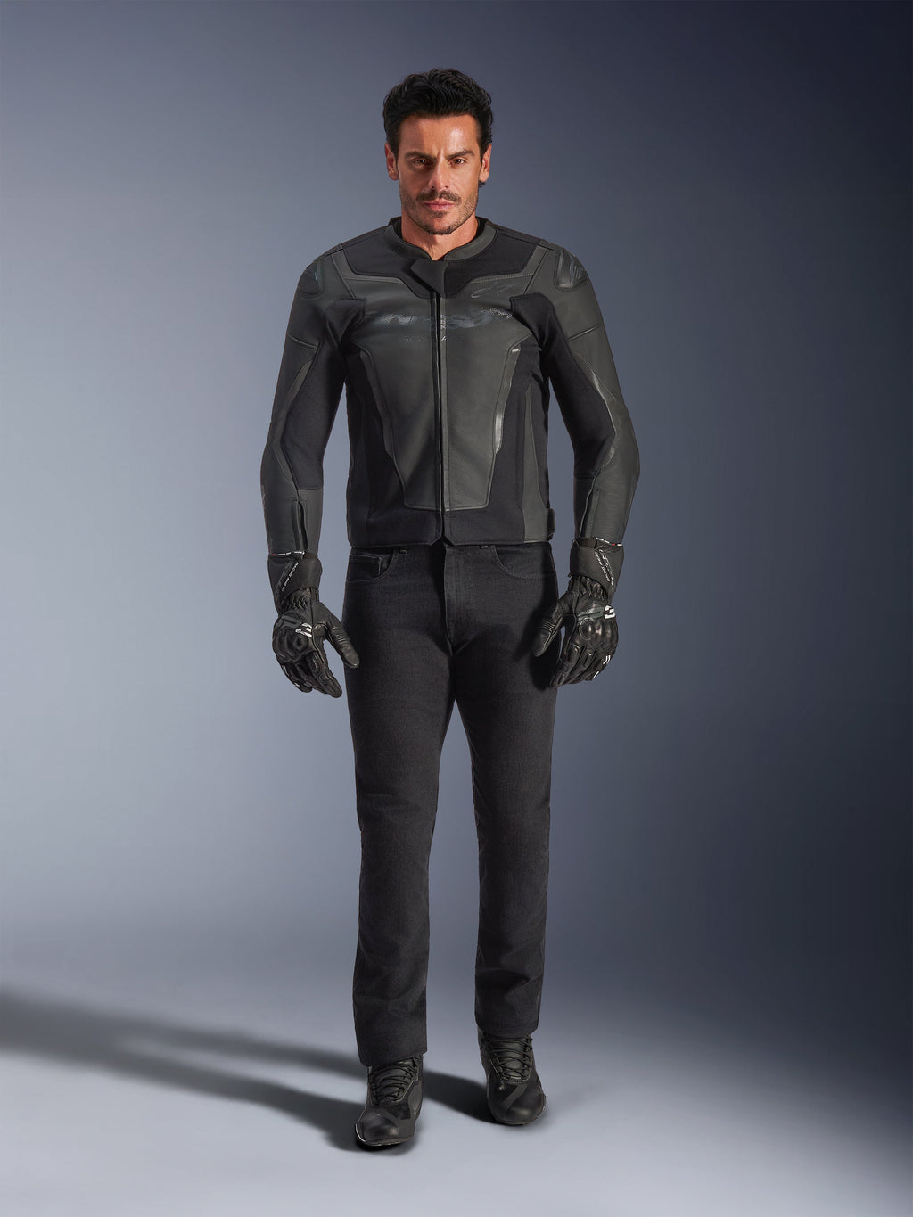 Veste en cuir Alpinestars GP Force V2 noire, portée par un motard avec un pantalon de moto, des gants et des bottes noirs, construction en cuir épurée de style sportif avec sliders d'épaules et protection intégrée.
