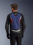 Rückansicht der Alpinestars GP Force V2 Lederjacke in Schwarz, Blau und Red Fluo, mit einem großen blauen Rückenpanel mit fluoreszierenden roten und hellblauen Akzenten, weißem Alpinestars-Logo und „Racing Ahead“-Schriftzug am unteren Rücken, getragen von einem Fahrer mit schwarzen Handschuhen und Hosen.