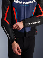 Alpinestars GP Force V2 Lederjacke in Schwarz, Blau und Fluo-Rot, Nahaufnahme einer Person beim Einstellen des Reißverschlusses am Ärmelbündchen, weiße Alpinestars-Logos auf Unterarm und Brust, strukturierte Rindslederkonstruktion für das sportliche Motorradfahren