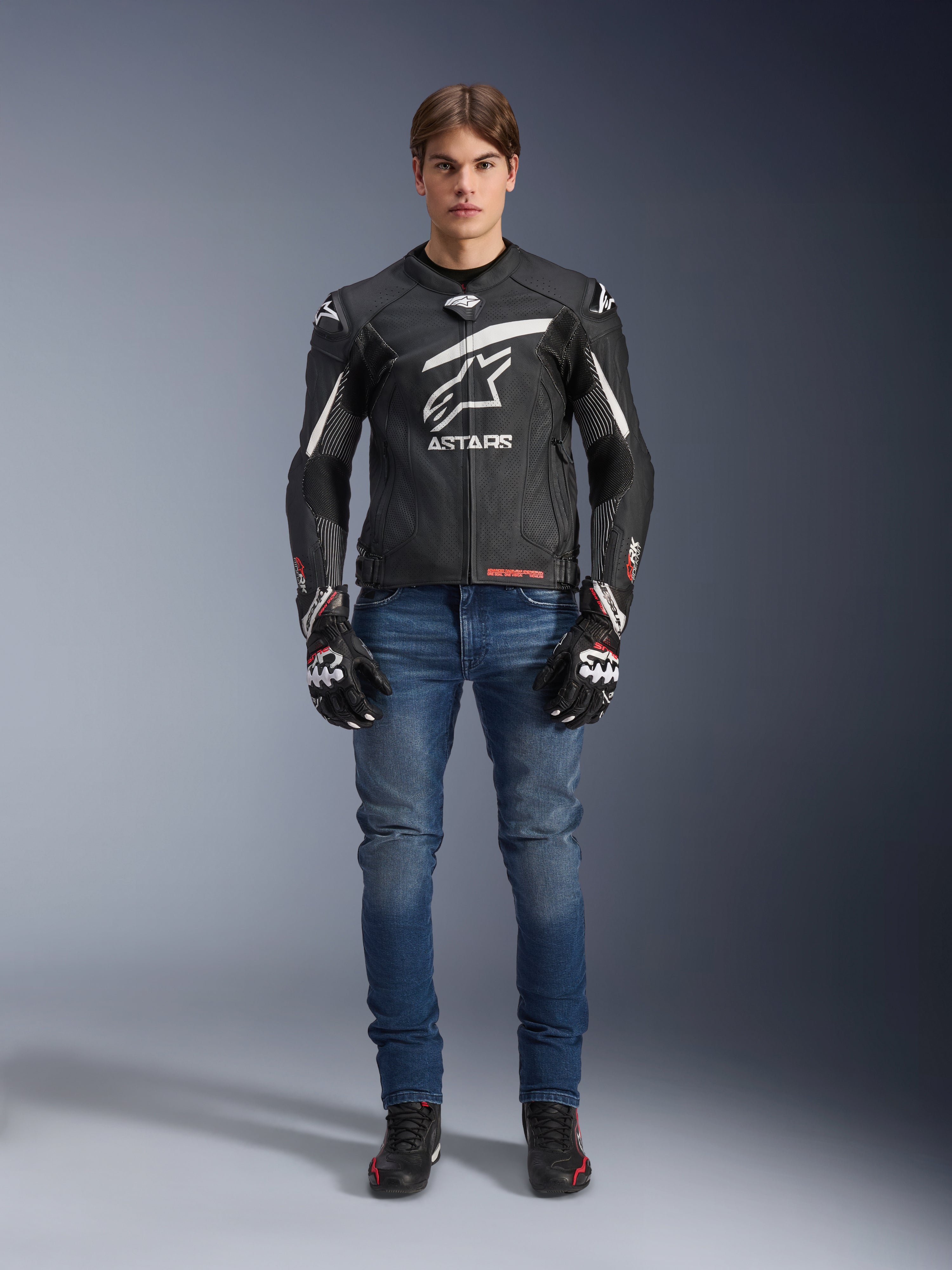 Alpinestars GP Plus R V4 Rideknit leren jas in zwart en wit, gedragen door een mannelijk model met blauwe motorjeans, zwarte motorhandschoenen en zwarte motorschoenen, voorzien van een groot wit Astars-logo op de borst, geperforeerde leren panelen en witte racestrepen op de mouwen, staand vooraanzicht.