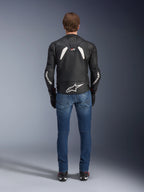 Achteraanzicht van de Alpinestars GP Plus R V4 Rideknit Leren Jas, zwart en wit, voorzien van een groot wit logo op de onderrug en branding op de mouwen, gedragen met een blauwe motorjeans en zwarte laarzen, wat de technische constructie en sportieve pasvorm benadrukt.
