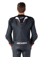 Rückansicht eines Fahrers in der Alpinestars Missile V3 Ignition Lederjacke, schwarzes Leder mit weißen und roten Akzenten, schmaler Sport-Aero-Höcker mit weißem Logo und 'alpinestars RACING AHEAD' Branding auf dem unteren Rücken, kombiniert mit passender schwarzer Leder-Rennhose und Handschuhen.