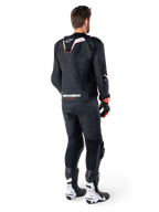 Rückansicht eines Fahrers in der Alpinestars Missile V3 Ignition Lederjacke in Schwarz, mit einem schmalen Sport-Aero-Höcker mit weißen und roten Akzenten, Alpinestars-Branding am unteren Rücken, kombiniert mit passender schwarzer Lederhose und schwarz-weißen Rennstiefeln, professionelles Sport-Fahreroutfit