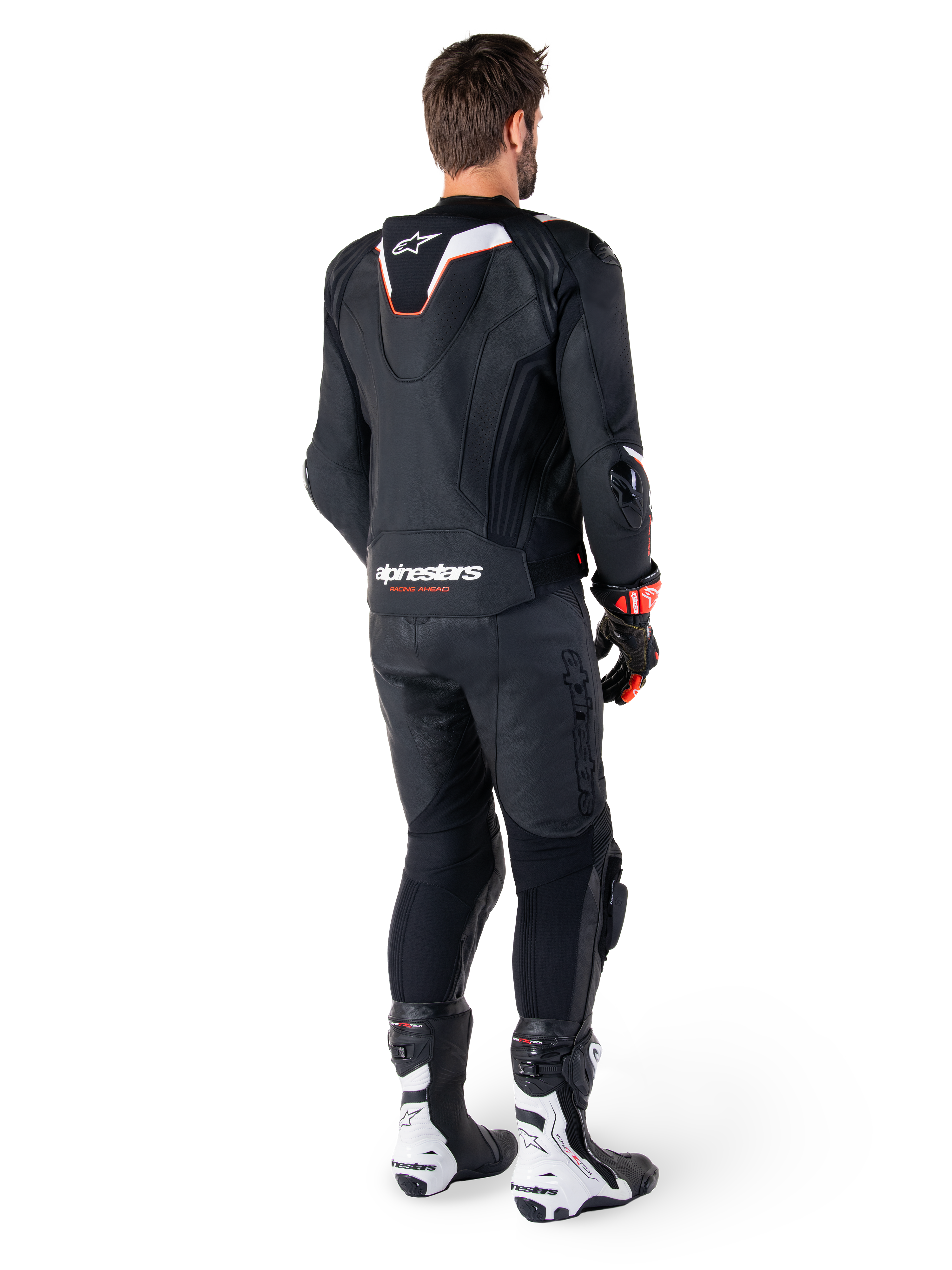Rückansicht eines Fahrers in der Alpinestars Missile V3 Ignition Lederjacke in Schwarz, mit einem schmalen Sport-Aero-Höcker mit weißen und roten Akzenten, Alpinestars-Branding am unteren Rücken, kombiniert mit passender schwarzer Lederhose und schwarz-weißen Rennstiefeln, professionelles Sport-Fahreroutfit