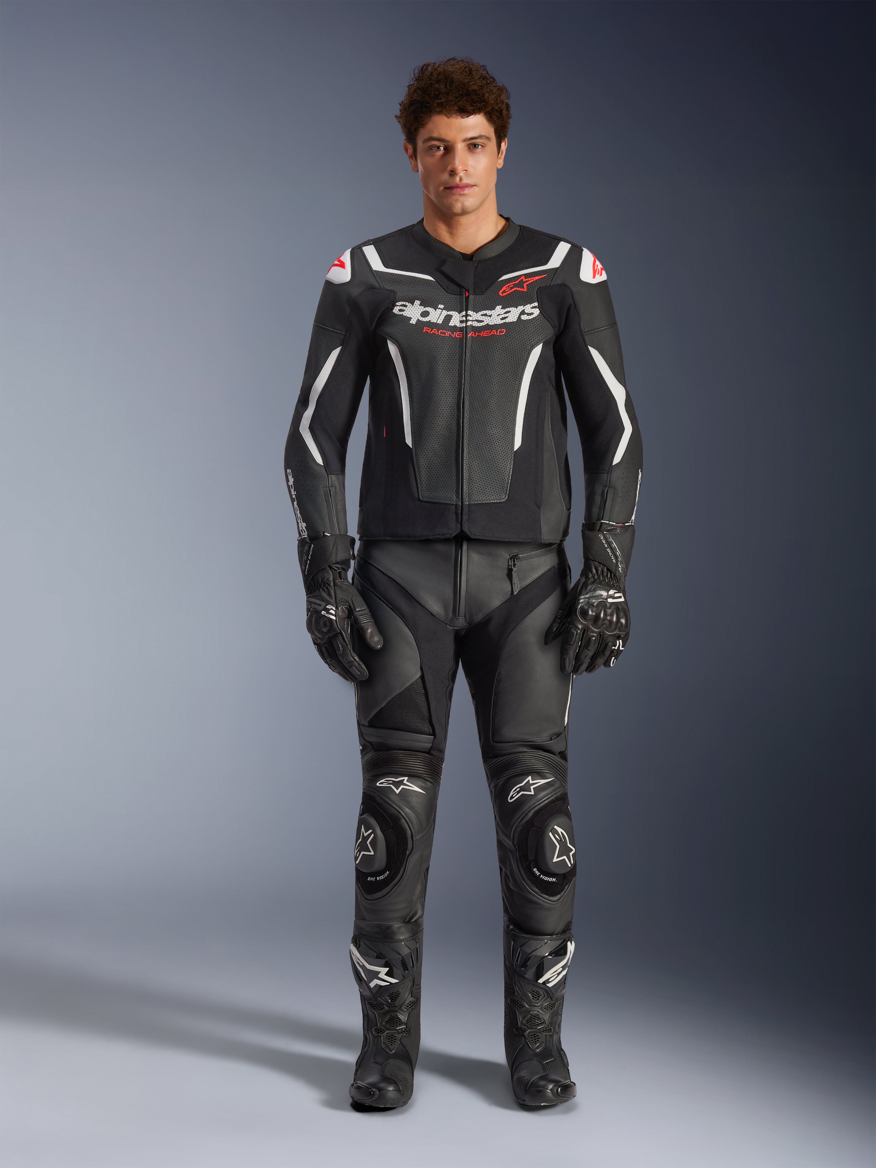 Mannelijk model draagt Alpinestars GP Force V2 Air leren jas, zwart en wit, gecombineerd met bijpassende leren broek, handschoenen en laarzen, sportief vooraanzicht, geperforeerde leren constructie met witte accenten en Alpinestars-logo's