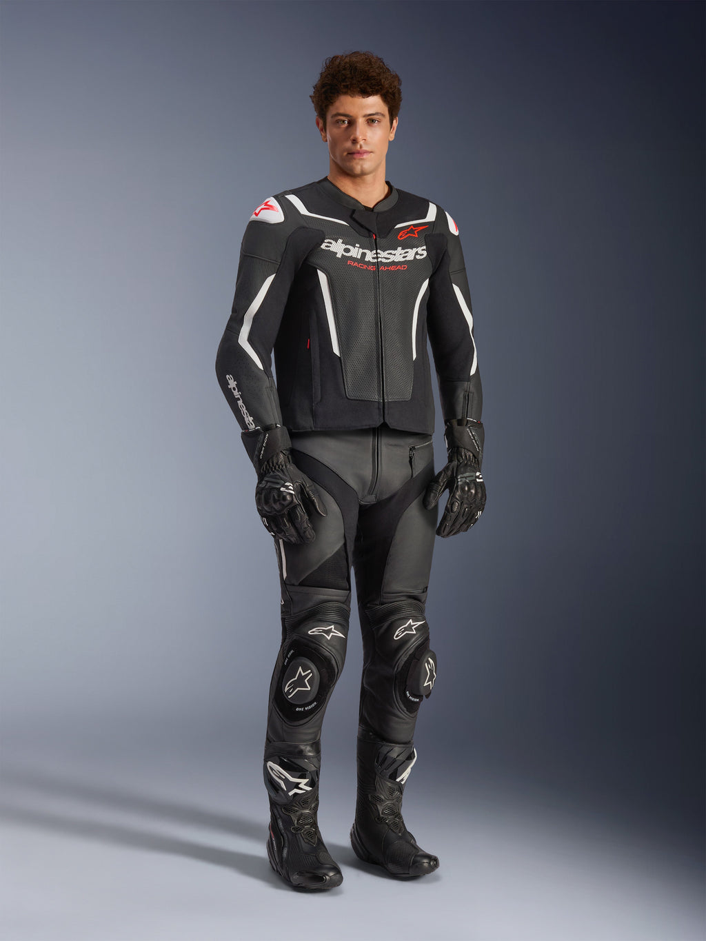 Alpinestars GP Force V2 Air Leren Jas in zwart en wit, gedragen door een mannelijke rijder met bijpassende zwarte racebroek, handschoenen en laarzen, voorzien van geperforeerde leren panelen, witte accenten en rood Alpinestars-logo op de borst, sportieve motoruitrusting