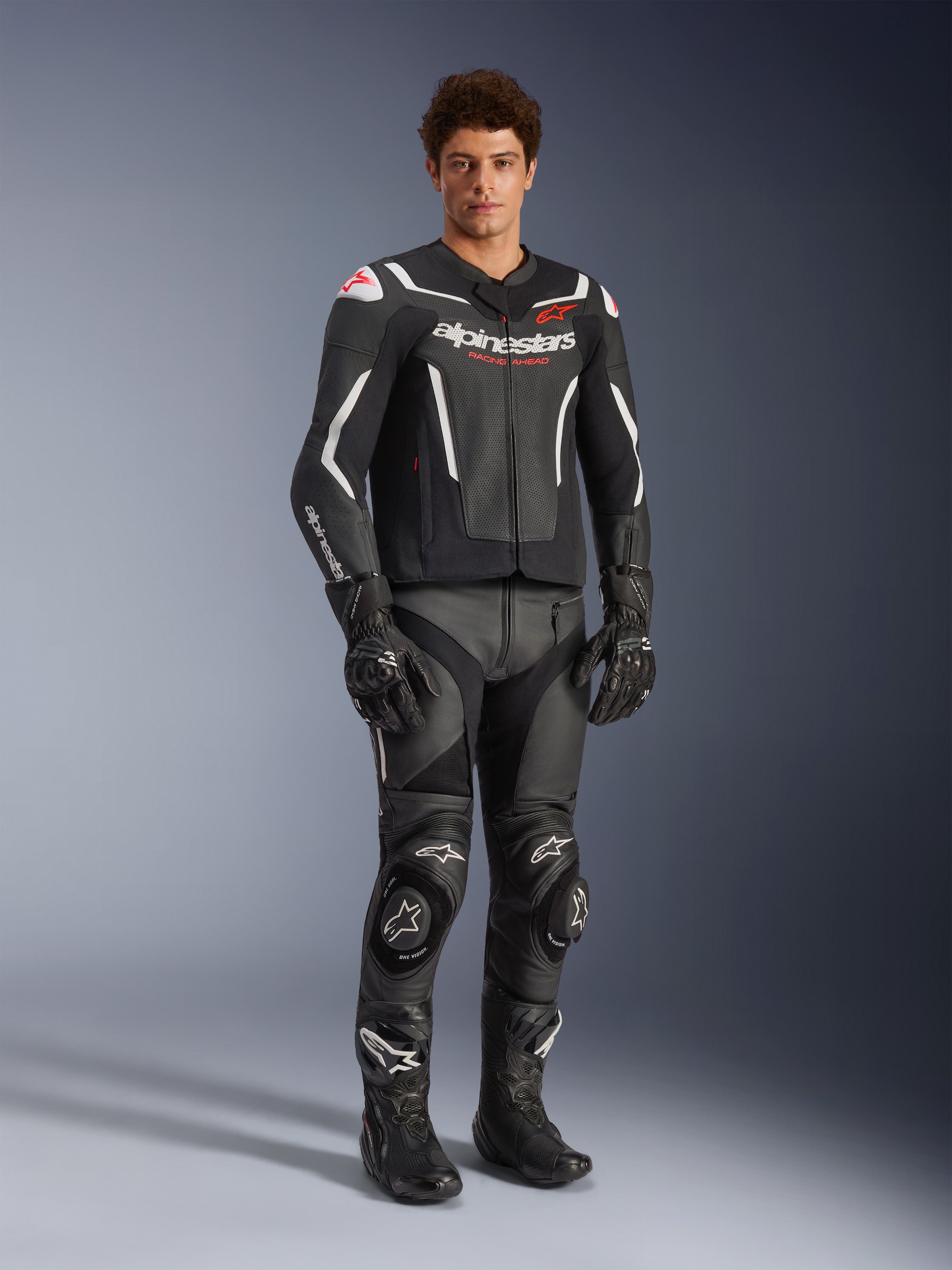 Alpinestars GP Force V2 Air Leren Jas in zwart en wit, gedragen door een mannelijke rijder met bijpassende zwarte racebroek, handschoenen en laarzen, voorzien van geperforeerde leren panelen, witte accenten en rood Alpinestars-logo op de borst, sportieve motoruitrusting
