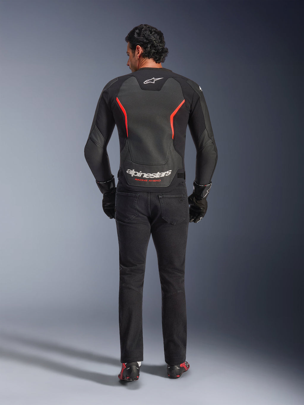 Alpinestars GP Force V2 Air Lederjacke in Schwarz und Rot Fluo, Rückansicht am Modell, mit perforierten Leder-Rückeneinsätzen, roten Akzenten und Alpinestars-Branding, kombiniert mit schwarzen Motorradjeans und Handschuhen für einen sportlichen Motorrad-Look.