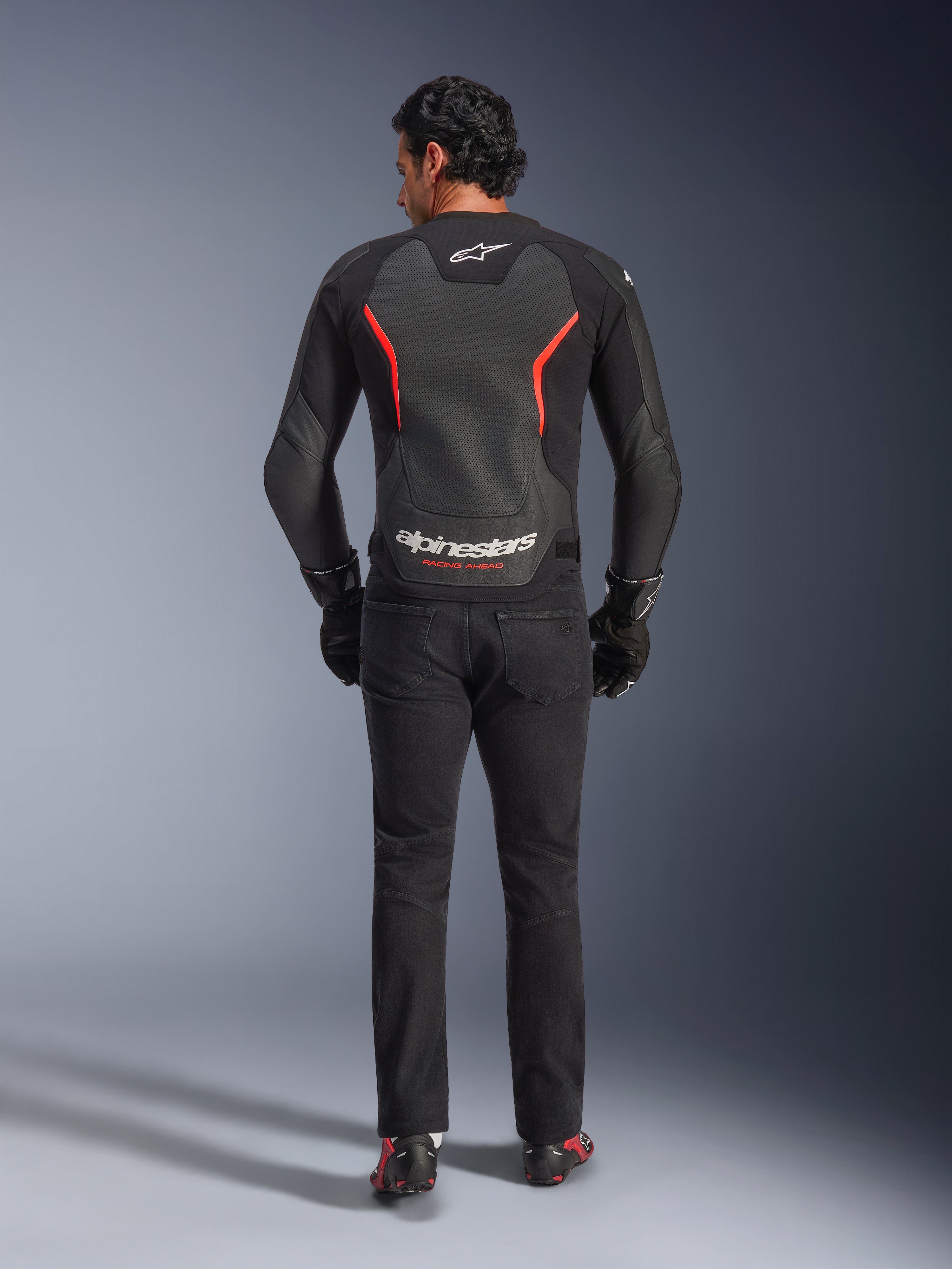 Alpinestars GP Force V2 Air Lederjacke in Schwarz und Rot Fluo, Rückansicht am Modell, mit perforierten Leder-Rückeneinsätzen, roten Akzenten und Alpinestars-Branding, kombiniert mit schwarzen Motorradjeans und Handschuhen für einen sportlichen Motorrad-Look.