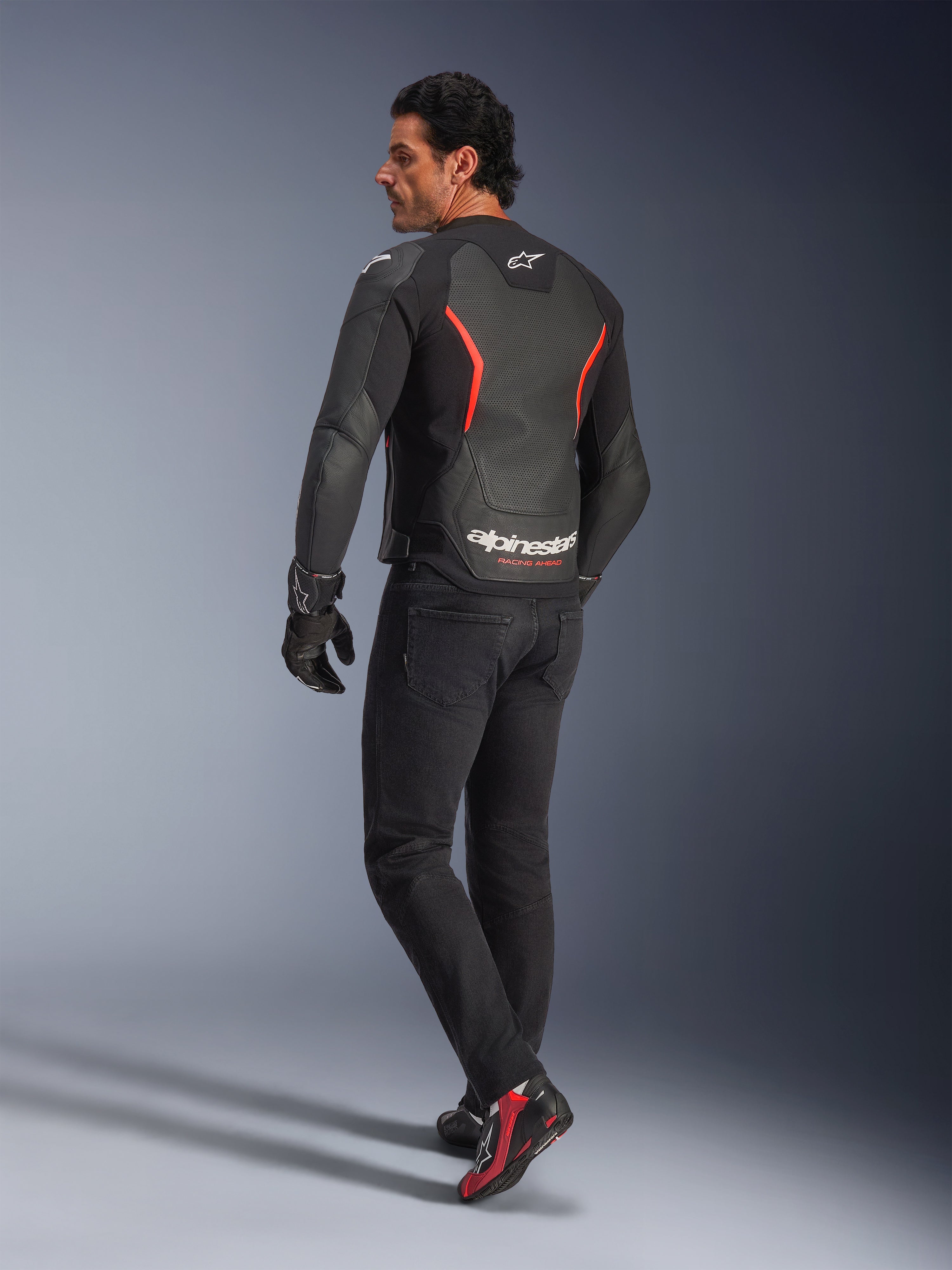 Männliches Model in der Rückansicht trägt die Alpinestars GP Force V2 Air Lederjacke in Schwarz-Rot-Fluo, mit umfassenden Perforationen am Rücken und markantem weißem Branding, kombiniert mit schwarzen Motorradjeans, Handschuhen und schwarz-roten Motorradstiefeln in einer Studioumgebung.