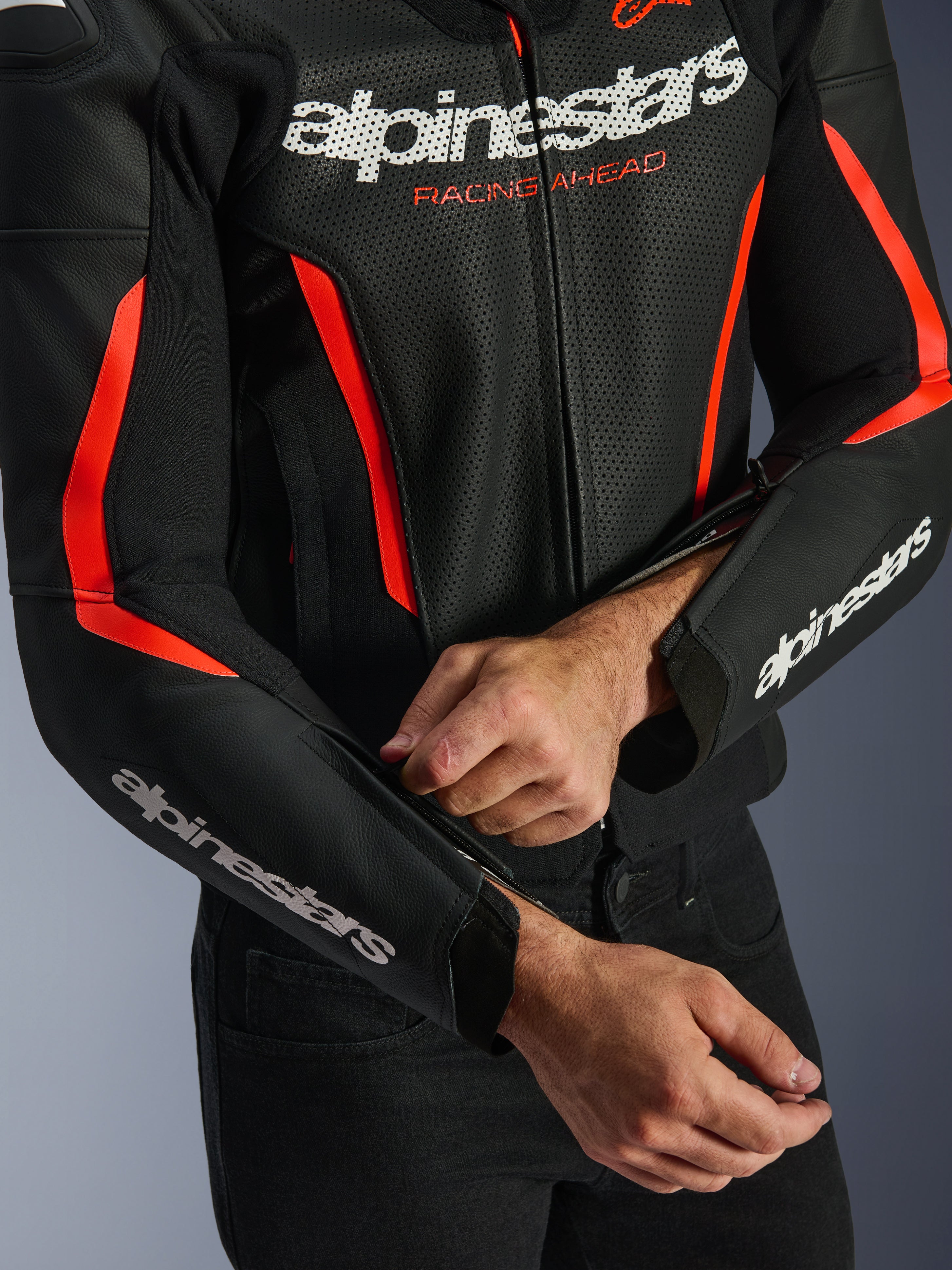 Alpinestars GP Force V2 Air Lederjacke in Schwarz und Rot Fluo, Nahaufnahme der perforierten Lederkonstruktion, weißes Branding auf Brust und Ärmeln, ein Fahrer passt den Unterarm-Reißverschluss für eine individuelle Passform an.