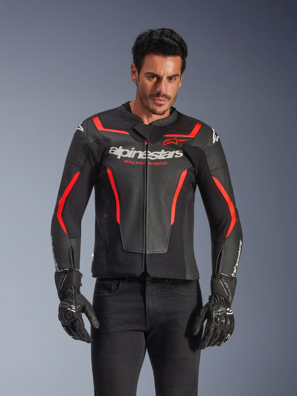 Alpinestars GP Force V2 Air Lederjacke, Motorrad-Rennjacke, Schwarz und Rot Fluo, hochwertige Lederkonstruktion mit großflächiger Perforation für optimale Belüftung, getragen von einem Model mit schwarzen Handschuhen und Hosen, mit markantem Branding und 'Racing Ahead'-Schriftzug auf der Brust
