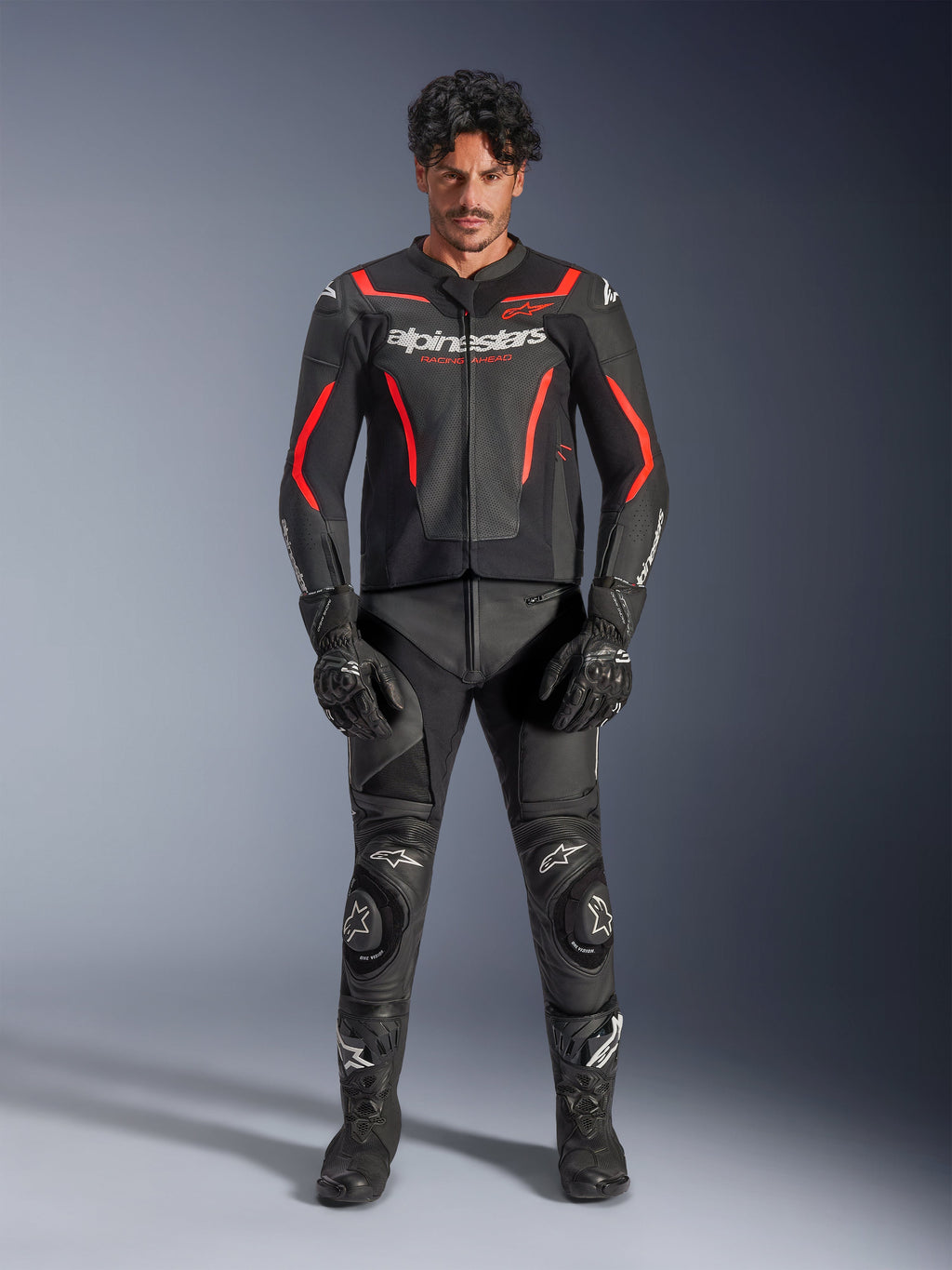 Männliches Model trägt die Alpinestars GP Force V2 Air Lederjacke in Schwarz und Red Fluo, mit perforierten Brusteinsätzen und weißem Branding, kombiniert mit schwarzer Motorrad-Lederhose, schwarzen Handschuhen und schwarzen Stiefeln für einen kompletten Racing-Look.