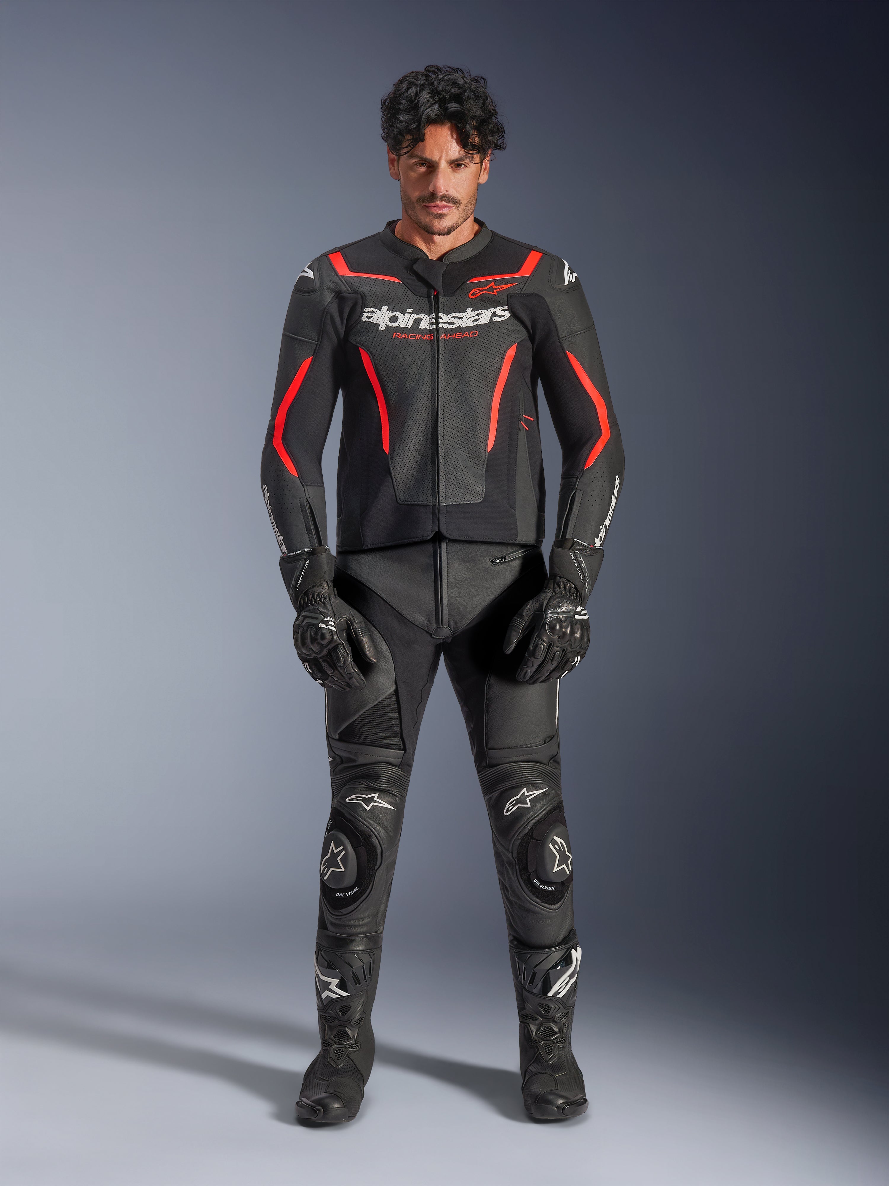Männliches Model trägt die Alpinestars GP Force V2 Air Lederjacke in Schwarz und Red Fluo, mit perforierten Brusteinsätzen und weißem Branding, kombiniert mit schwarzer Motorrad-Lederhose, schwarzen Handschuhen und schwarzen Stiefeln für einen kompletten Racing-Look.
