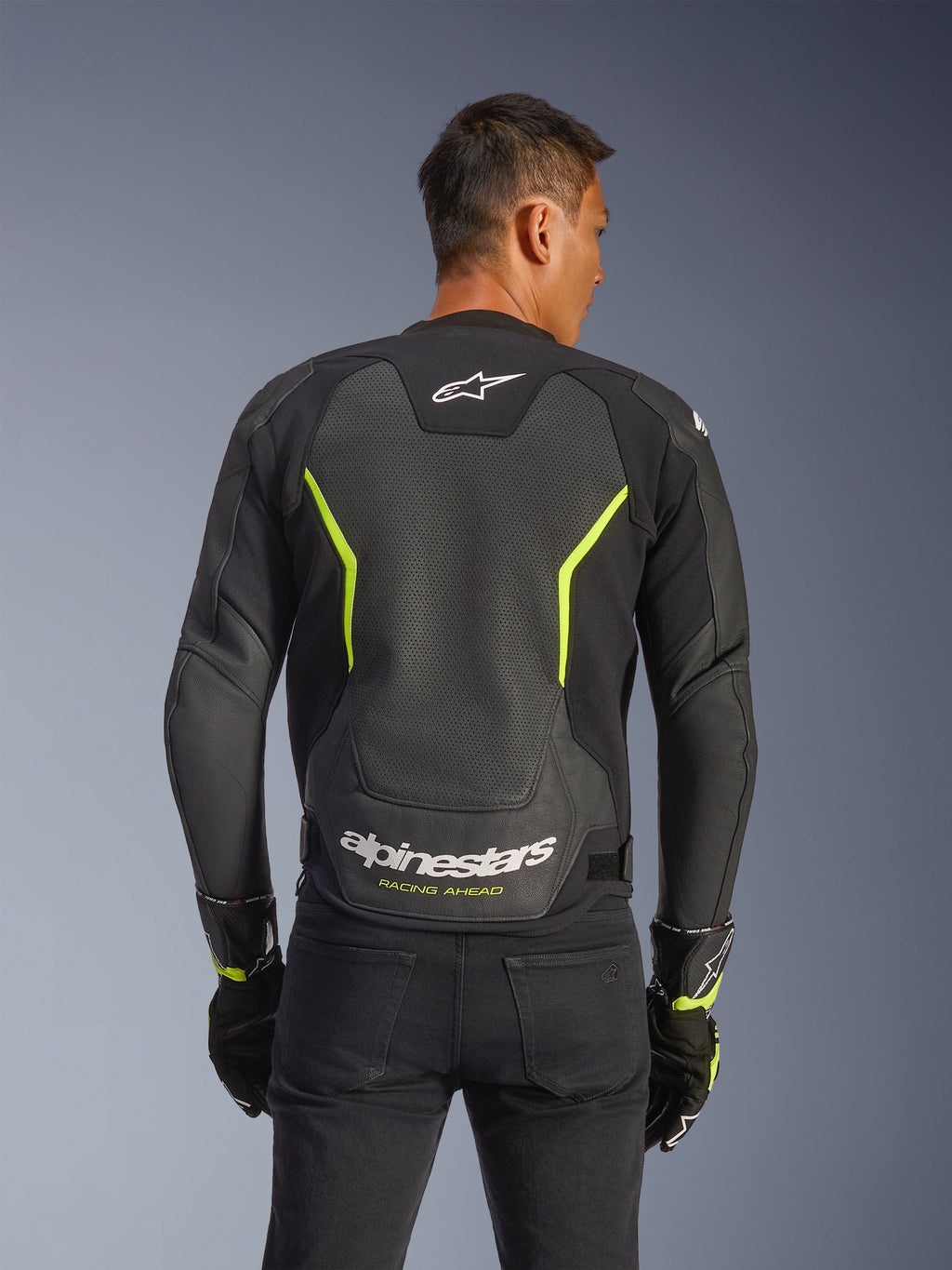 Blouson en cuir Alpinestars GP Force V2 Air, noir et jaune fluo, vue de dos d'un pilote montrant de larges panneaux en cuir perforé pour la circulation de l'air, marquage Alpinestars blanc et texte « Racing Ahead » jaune au bas du dos, équipement de moto sportif pour la route.