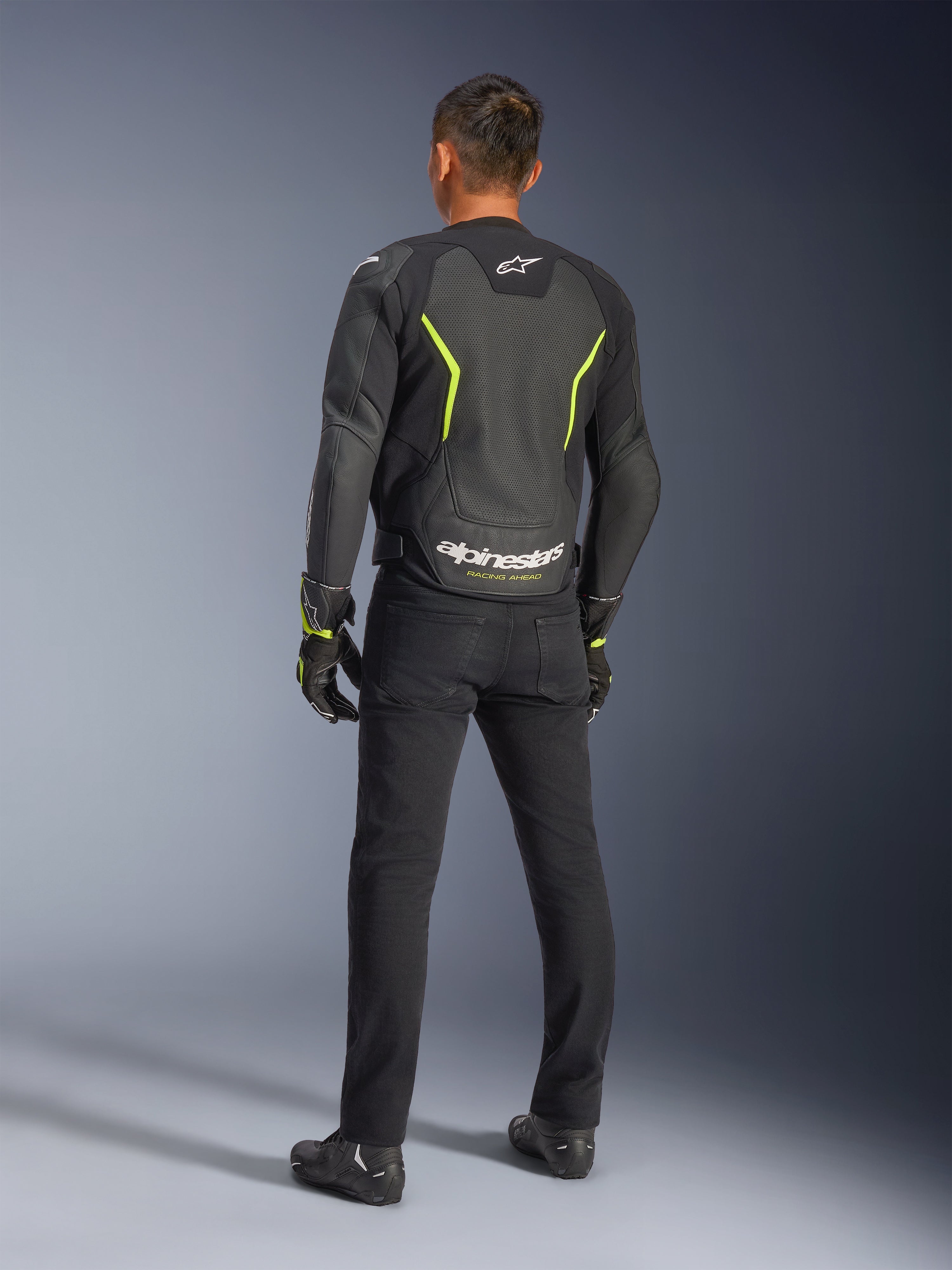 Alpinestars GP Force V2 Air Leather Jacket, noir et jaune fluo, vue arrière d'un pilote portant le blouson en cuir perforé, doté d'accents jaunes angulaires et d'un logo Alpinestars blanc avec 'RACING AHEAD' en bas du dos, avec pantalon de moto et bottes noirs.