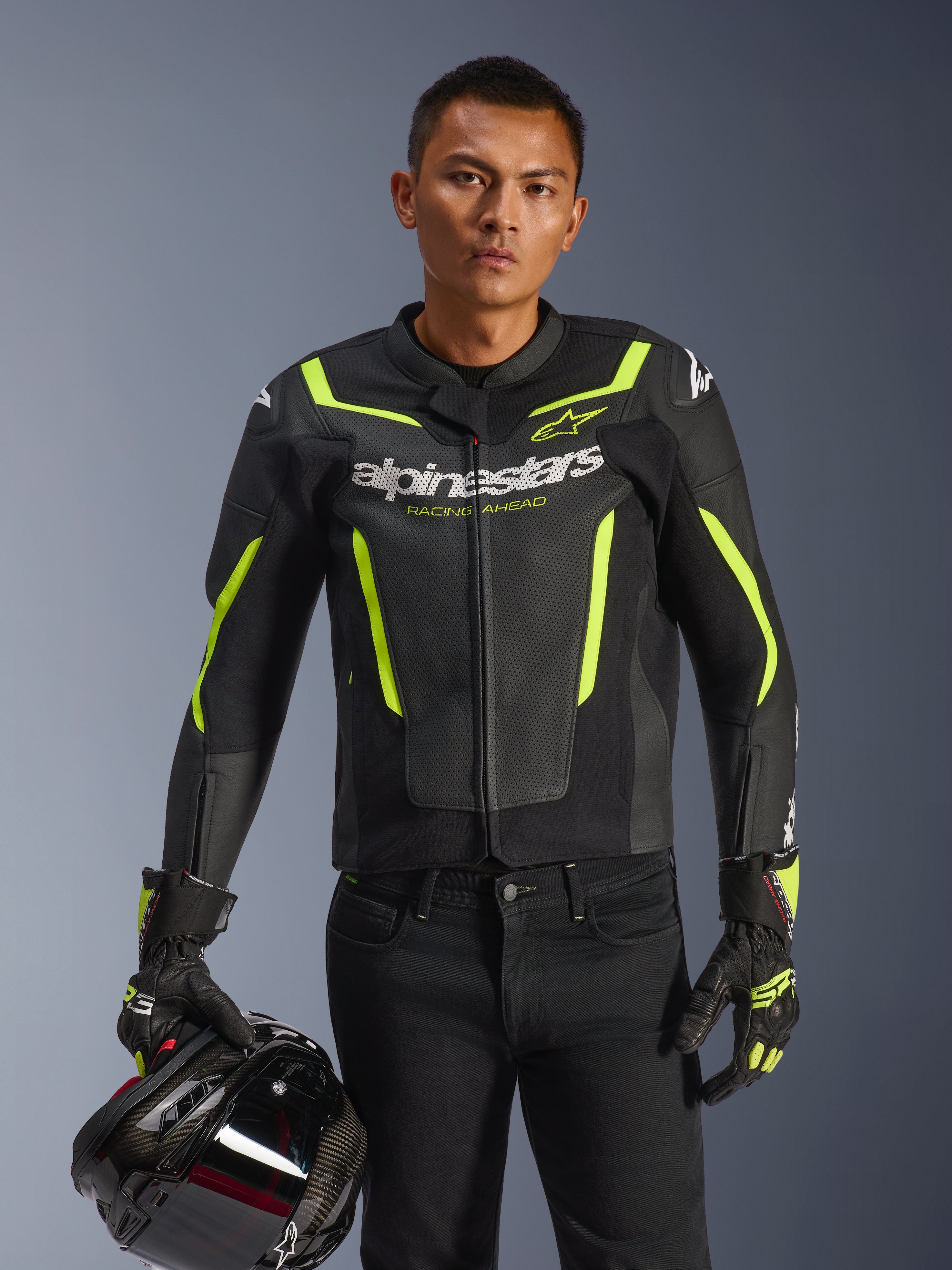 Pilote portant la veste en cuir Alpinestars GP Force V2 Air en noir et jaune fluo, tenant un casque de moto noir, cuir perforé avec logo Alpinestars blanc et détails jaune fluo, gants de course assortis et pantalon noir