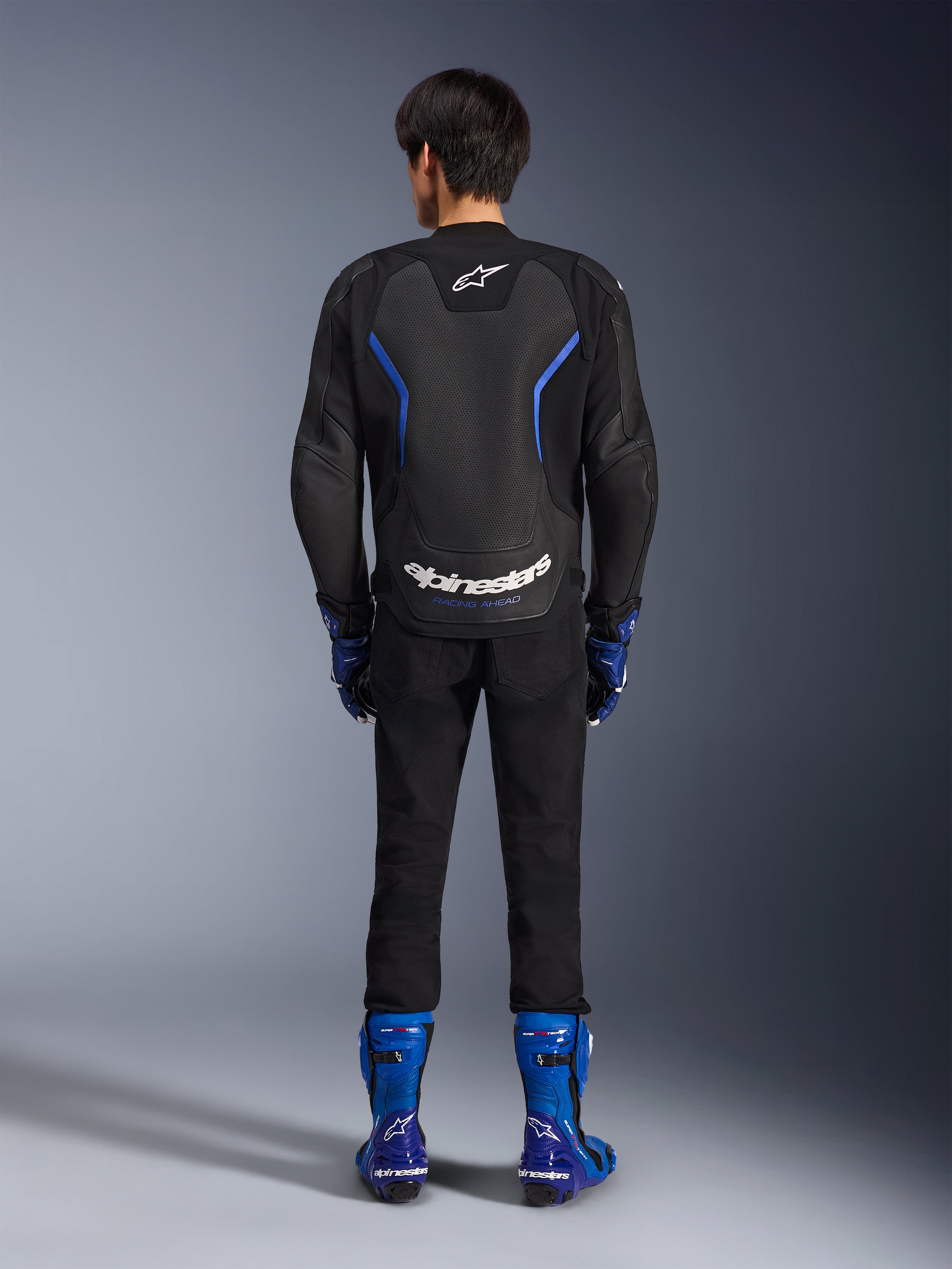 Blouson en cuir Alpinestars GP Force V2 Airflow, Noir Bleu Métallique, vue arrière montrant des panneaux en cuir perforé et un marquage blanc, porté avec des bottes de course bleues et un pantalon noir, équipement de course moto sportive