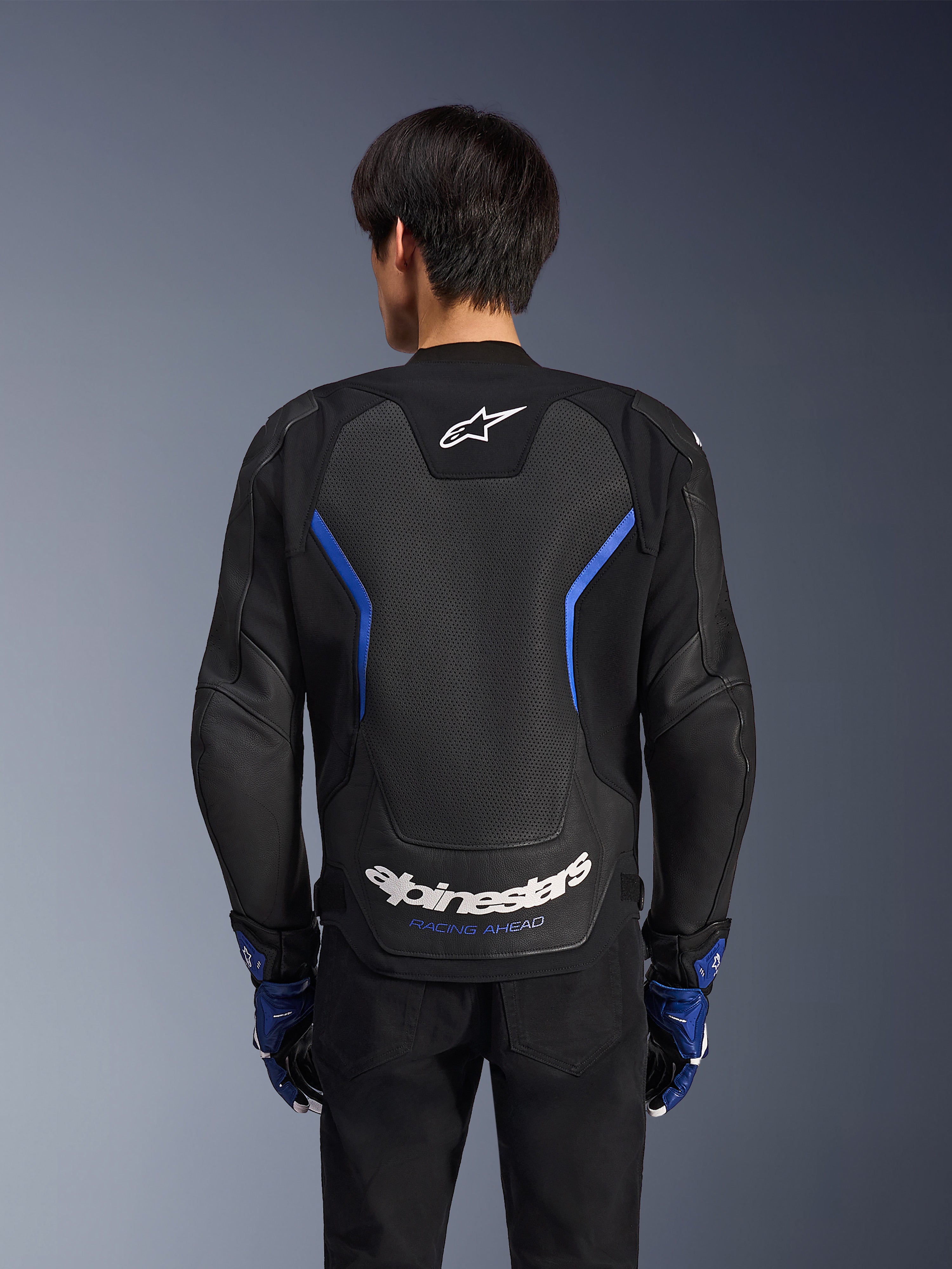 Blouson en cuir Alpinestars GP Force V2 Airflow, Noir Bleu Métallisé, vue arrière, doté d'un grand panneau perforé pour la ventilation, d'accents bleus et du logo Alpinestars blanc, porté avec des gants bleus assortis et un pantalon noir pour la conduite sportive.