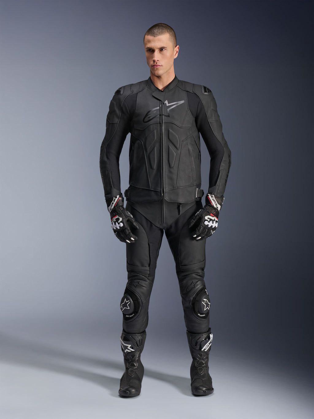 Alpinestars Dusk Lederjacke, Schwarz, getragen von einem männlichen Model als Teil eines kompletten, vom Rennsport inspirierten Outfits mit passender Lederhose, Handschuhen und Stiefeln, schlankes ergonomisches Design mit Protektoren an den Schultern und Markenlogo auf der Brust.