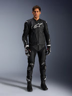 Mannelijk model draagt de Alpinestars Dusk leren jas in zwart en wit, met opvallende witte logo's op de borst en schouders, gecombineerd met een bijpassende zwarte leren motorbroek, zwart-witte handschoenen en zwarte laarzen, complete sportieve race-outfit voor weggebruik.