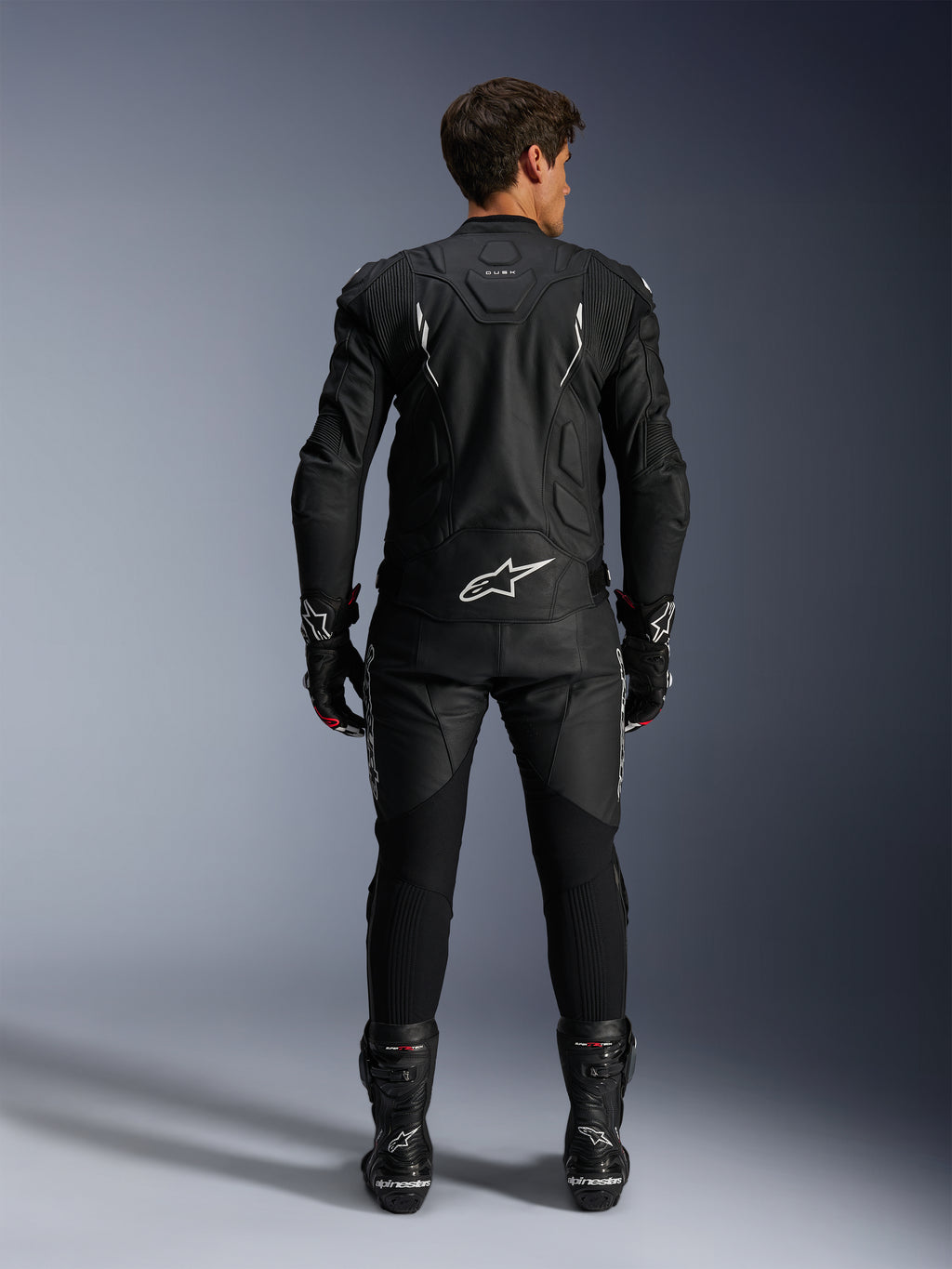 Alpinestars motorrijder van achteren gezien, Dusk Leather Jacket en Missile V3 Leather Pants, volledig zwart leren pak met witte accenten, jas met 'Dusk' branding en Alpinestars logo op de rug, gecombineerd met zwarte racelaarzen en handschoenen, gecoördineerde motorsport outfit voor wegraces