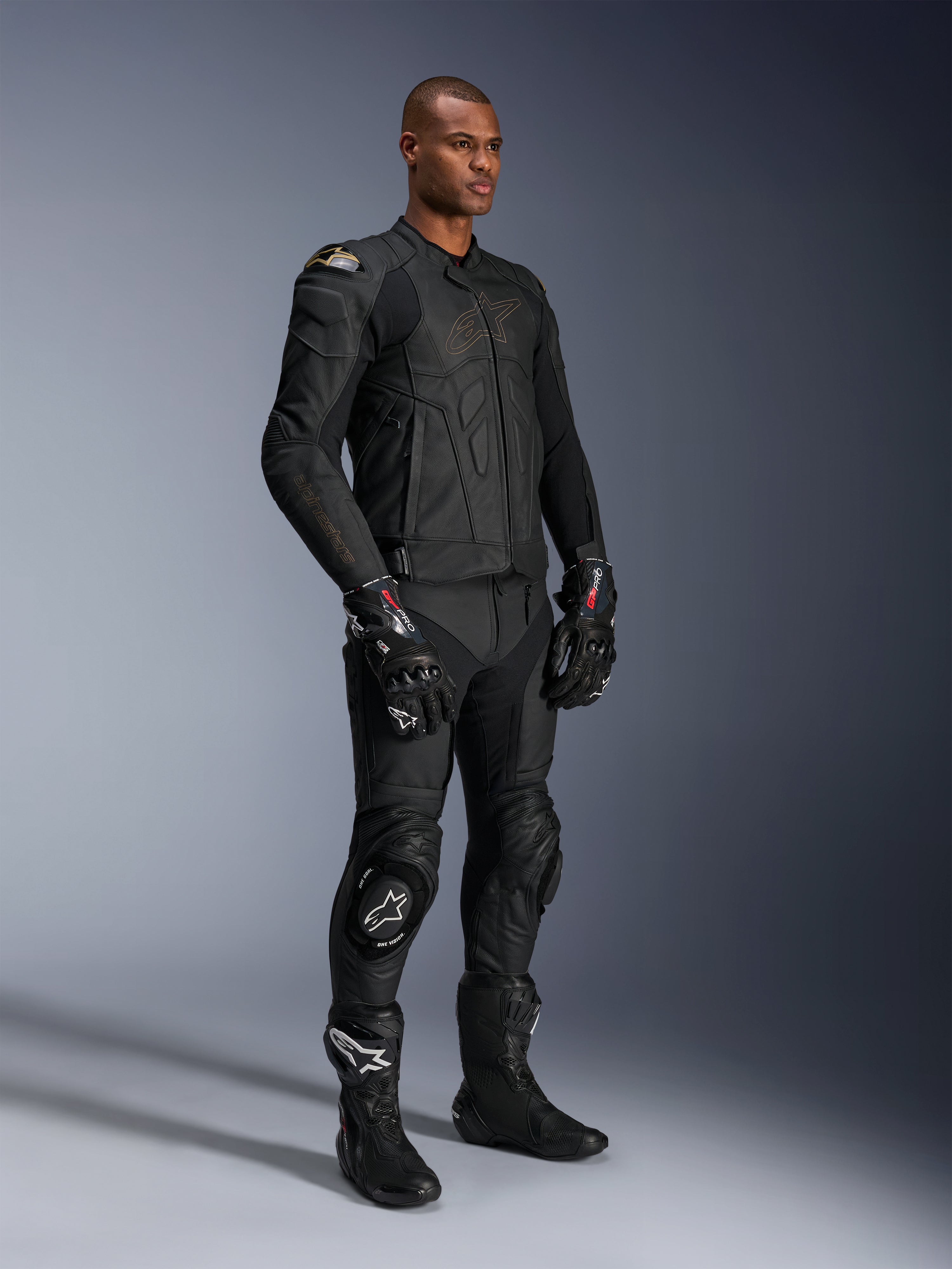 Mannelijk model in Alpinestars Missile V3 leren broek en bijpassende zwarte leren jas met gouden accenten, zwarte GP Pro racehandschoenen en zwarte racelaarzen, complete motorrace-uitrusting voor weggebruik, studio-opname.