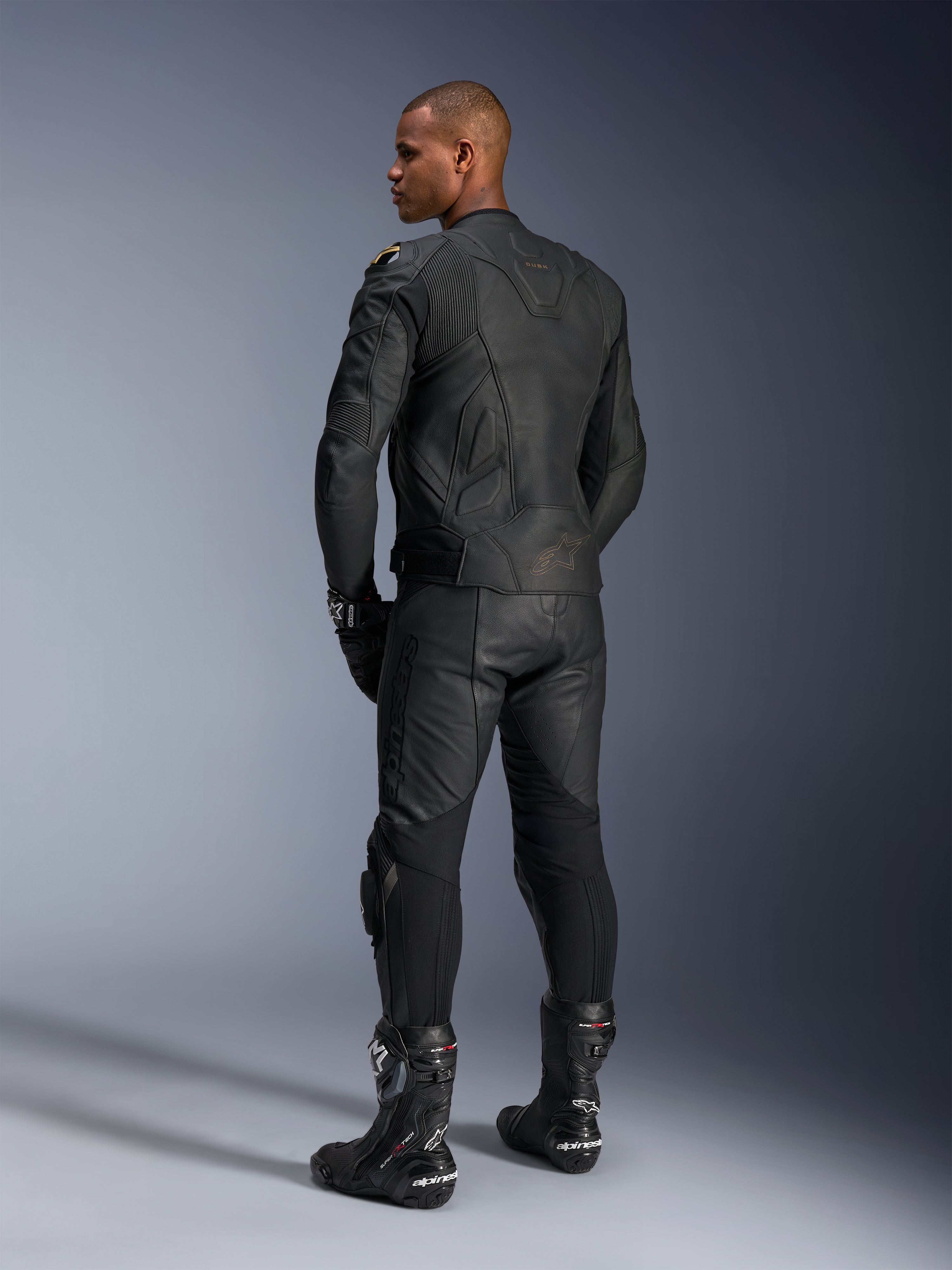 Alpinestars Missile V3 leren broek en Dusk leren jas, zwart met gouden accenten, achteraanzicht van de rijder, met logo's in reliëf en accordeon stretchpanelen, gedragen met zwarte Supertech R laarzen en handschoenen, professionele motorsportkleding.