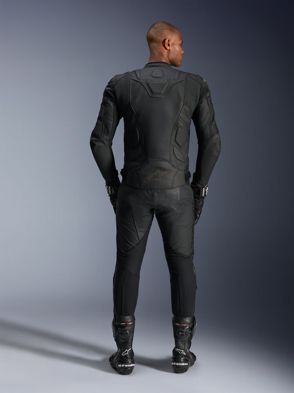 Alpinestars Dusk Leren Jas, Black Laser, achteraanzicht op een motorrijder, gedragen met bijpassende leren motorbroek, racehandschoenen en laarzen, voorzien van ergonomische 3D multi-panel constructie en beschermende leren panelen.